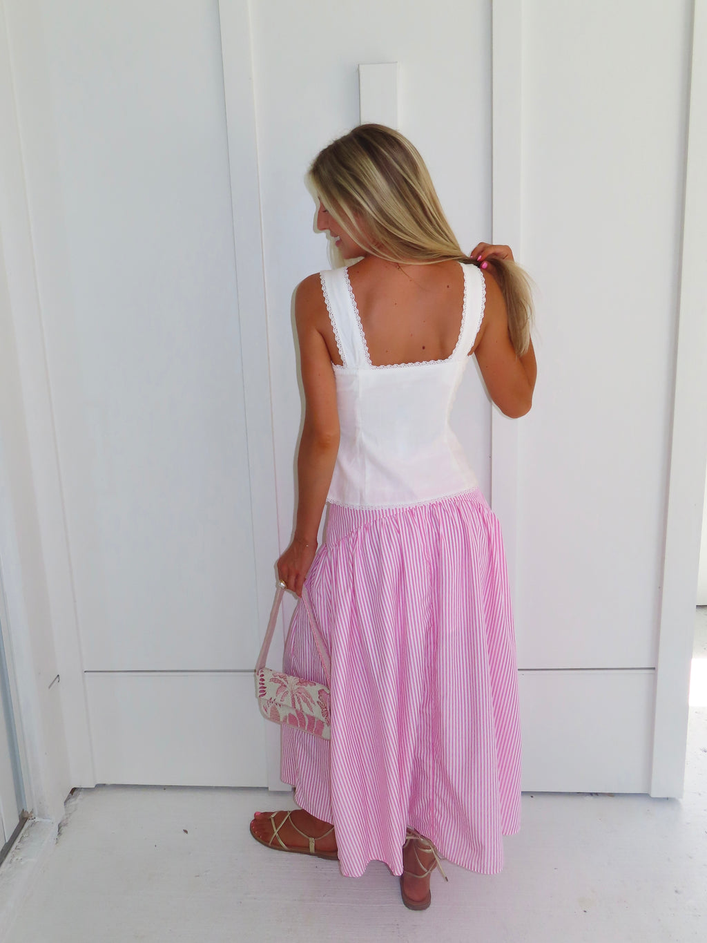 A woman wearing a pink and white striped maxi skirt with a white tank top and gold sandals, holding a pink and white purse and standing against a white paneled wall.