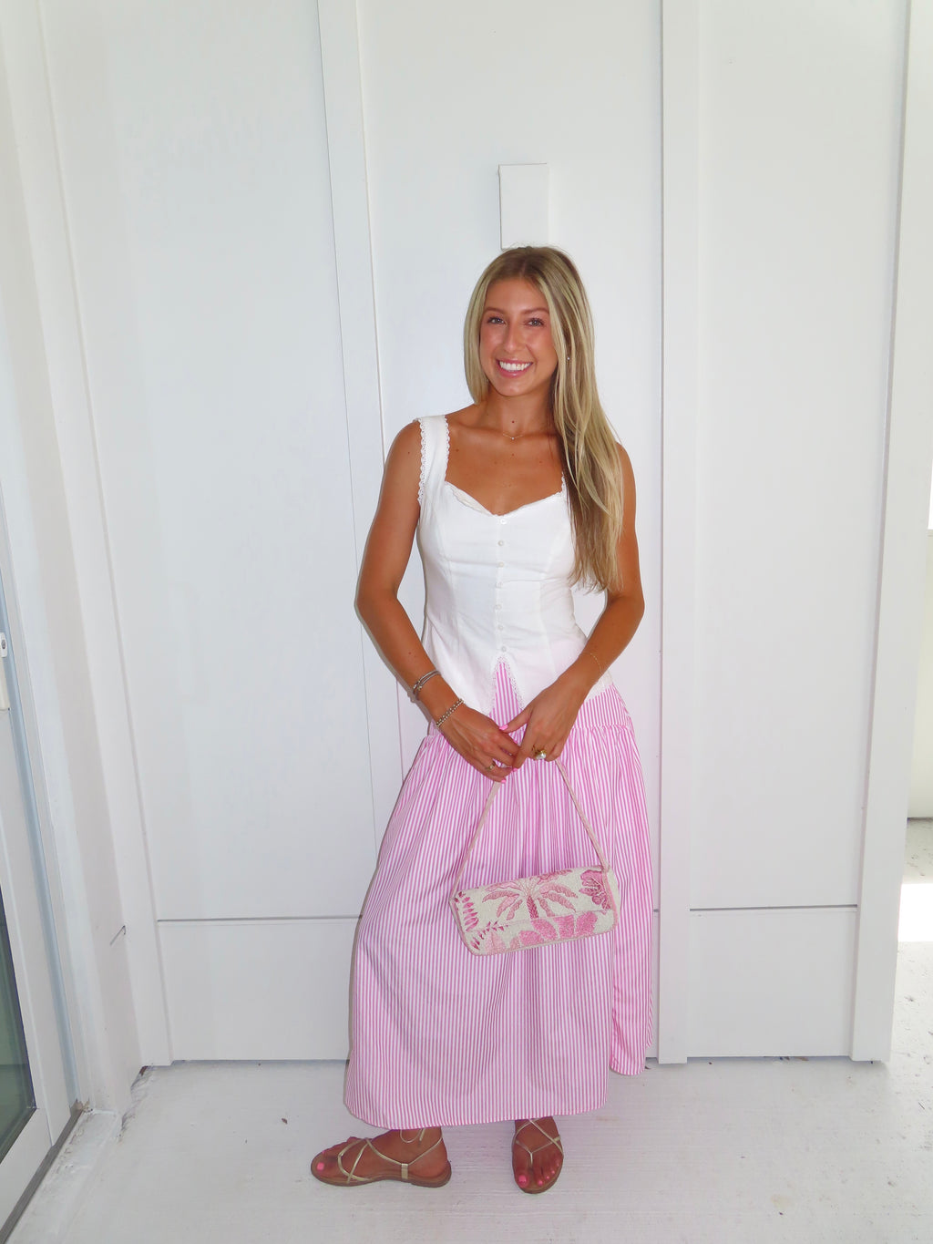 A woman wearing a pink and white striped maxi skirt with a white tank top and gold sandals, holding a pink and white purse and standing against a white paneled wall.