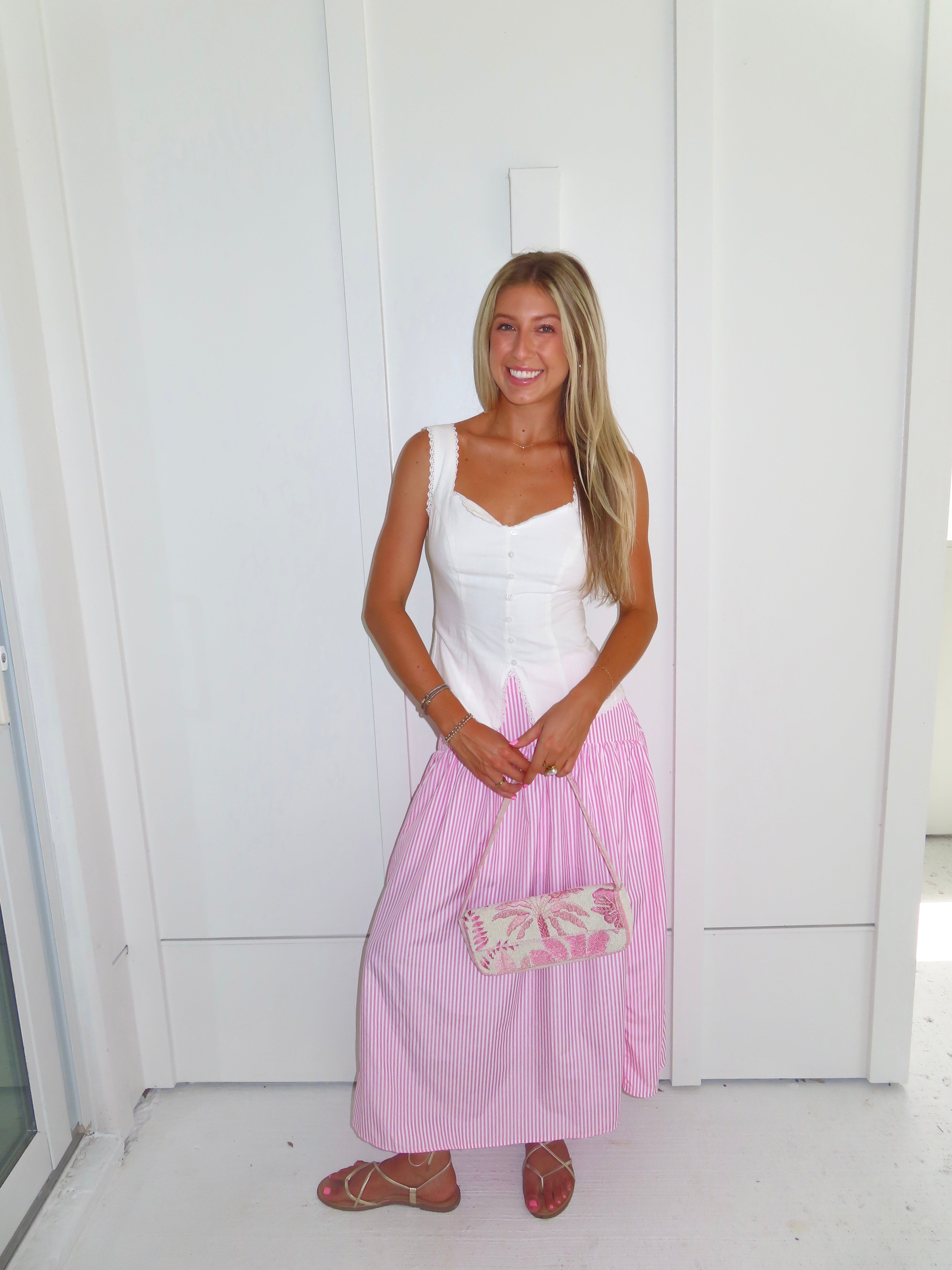 A woman wearing a pink and white striped maxi skirt with a white tank top and gold sandals, holding a pink and white purse and standing against a white paneled wall.