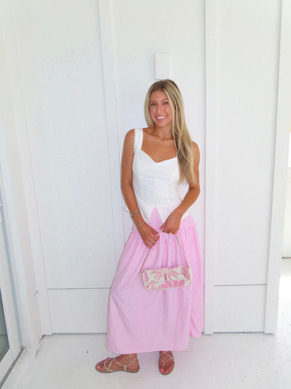 A woman wearing a pink and white striped maxi skirt with a white tank top and gold sandals, holding a pink and white purse and standing against a white paneled wall.