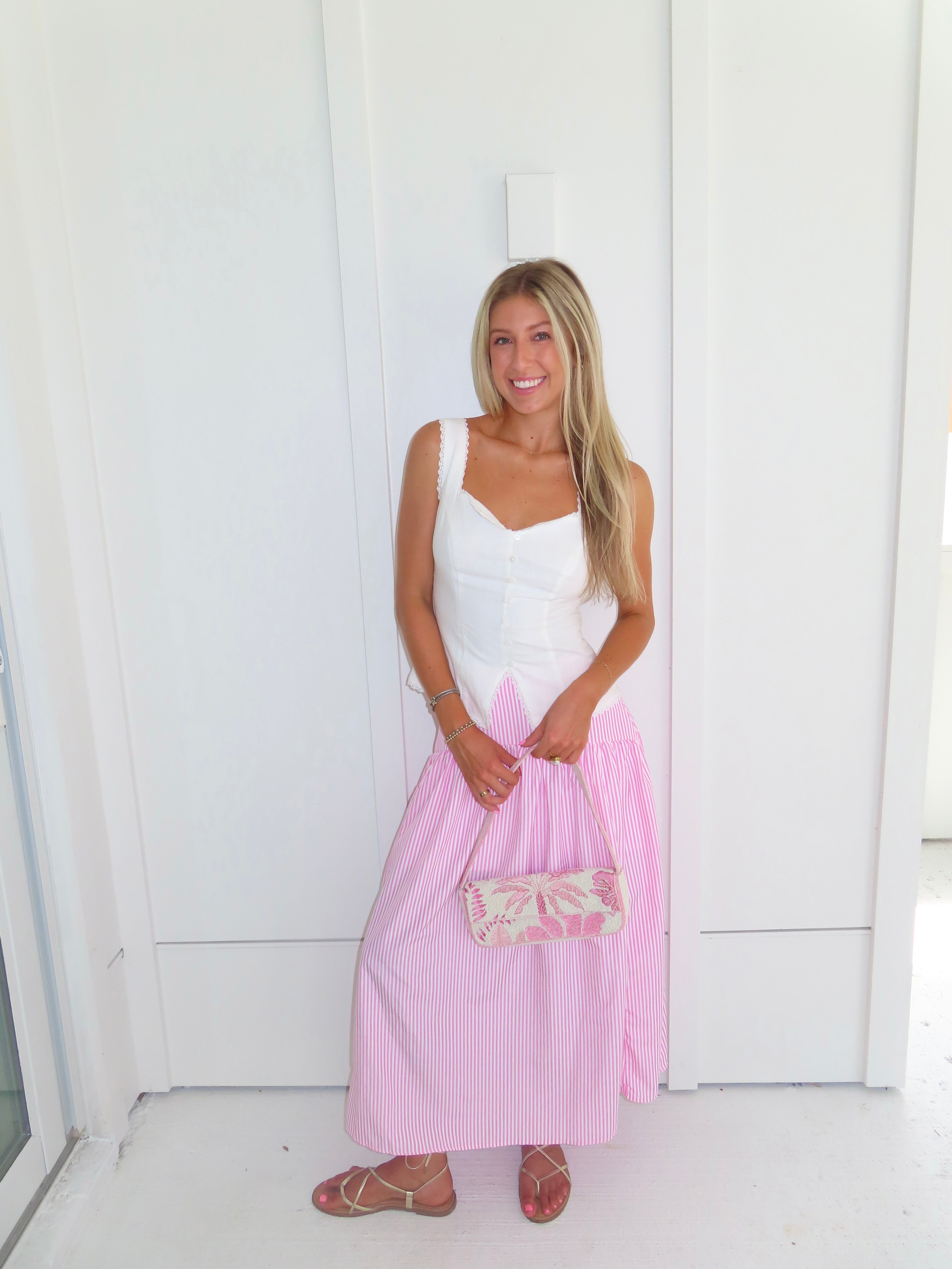 A woman wearing a pink and white striped maxi skirt with a white tank top and gold sandals, holding a pink and white purse and standing against a white paneled wall.