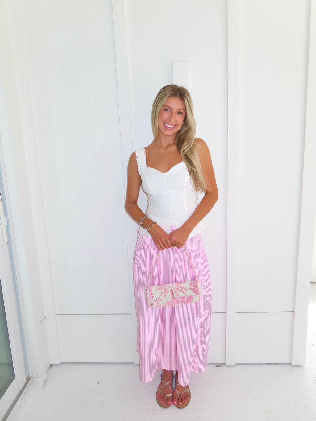 A woman wearing a pink and white striped maxi skirt with a white tank top and gold sandals, holding a pink and white purse and standing against a white paneled wall.