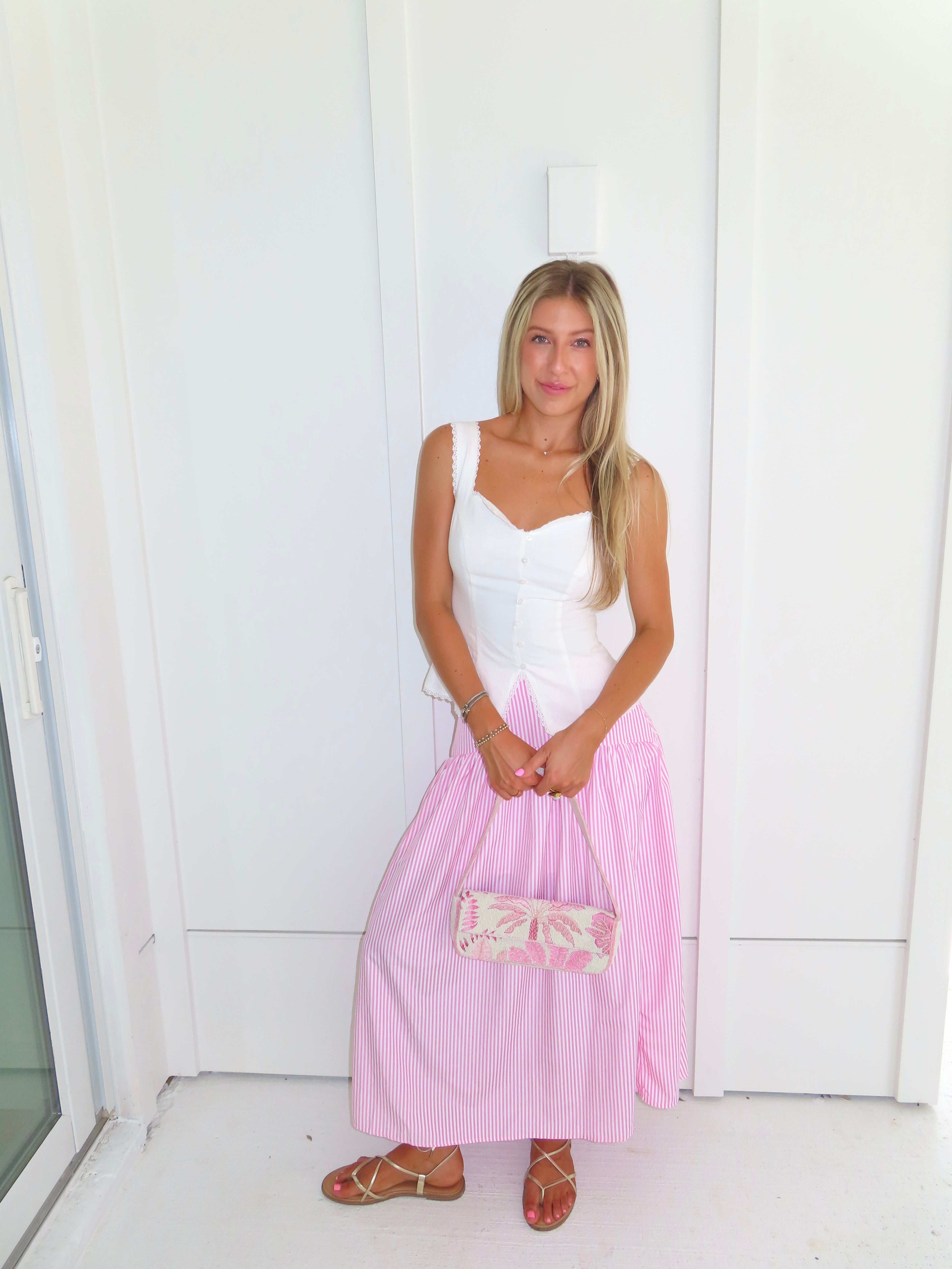 A woman wearing a pink and white striped maxi skirt with a white tank top and gold sandals, holding a pink and white purse and standing against a white paneled wall.
