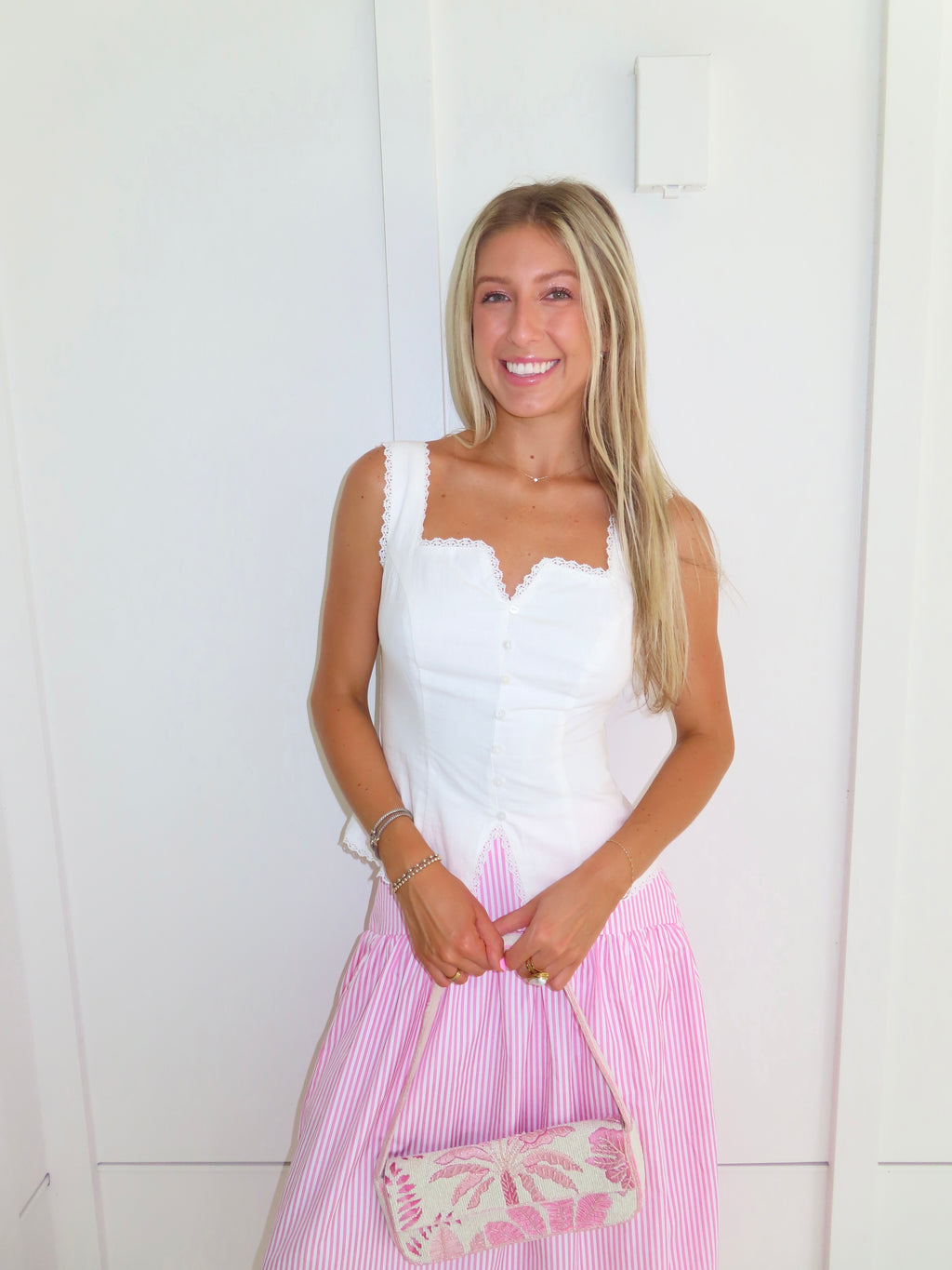 A woman wearing a pink and white striped maxi skirt with a white tank top, holding a pink and white purse and standing against a white paneled wall.
