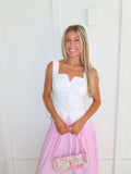 A woman wearing a pink and white striped maxi skirt with a white tank top, holding a pink and white purse and standing against a white paneled wall.