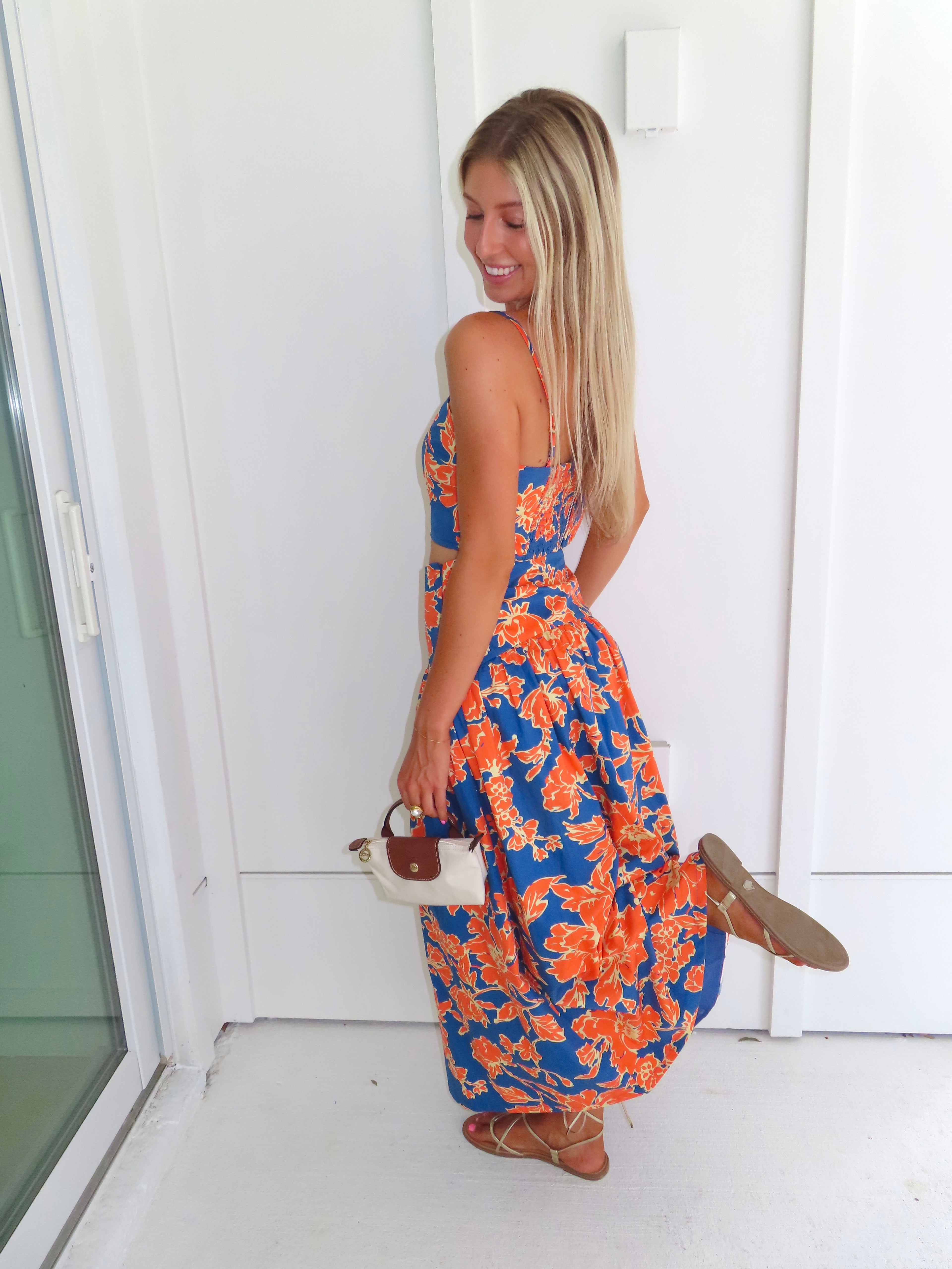 A woman posed wearing a sleeveless maxi skirt set with a bold orange and blue floral print. She is holding a small beige handbag with a brown leather detail and wearing gold strappy sandals.