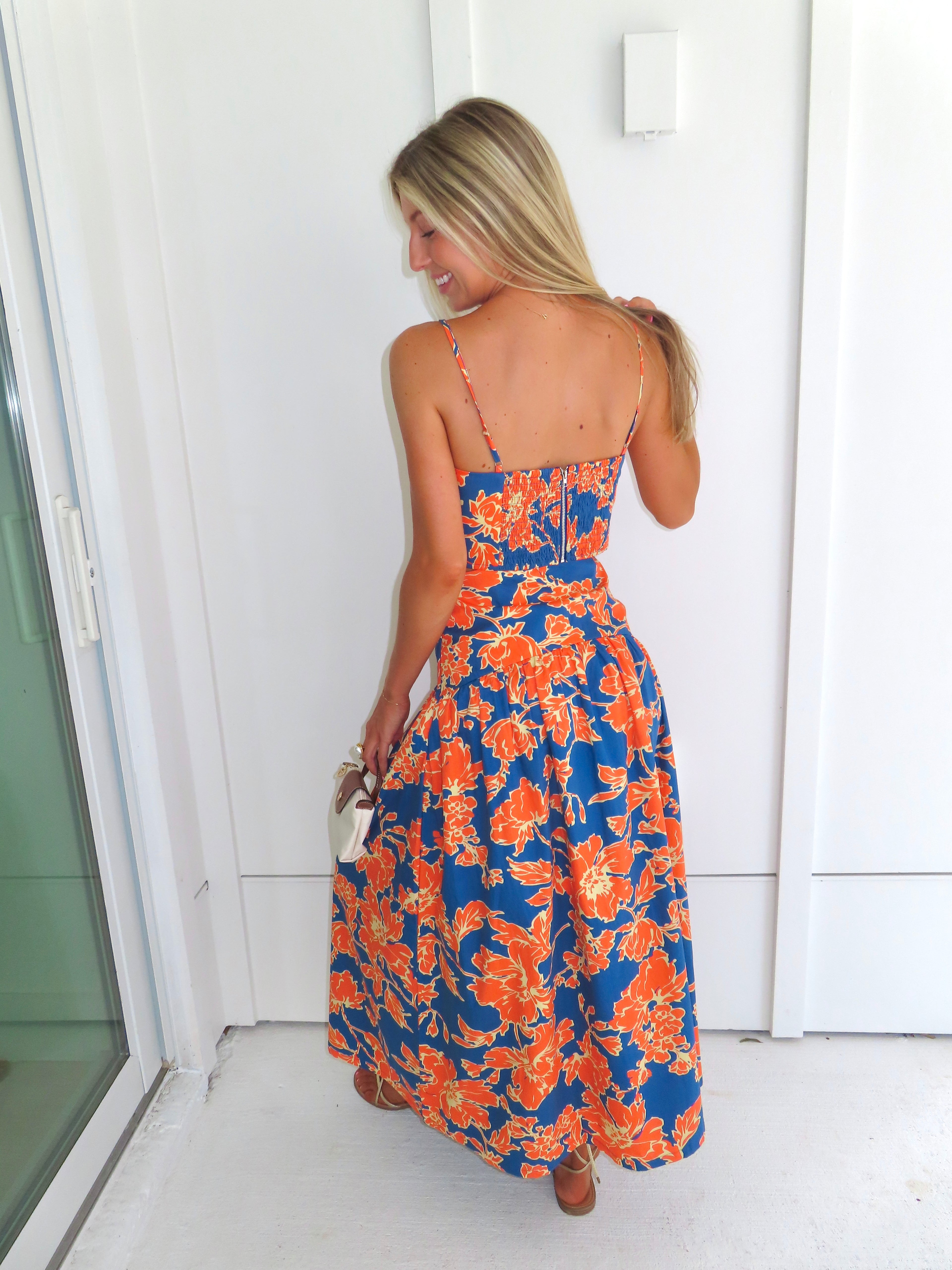A woman posed wearing a sleeveless maxi skirt set with a bold orange and blue floral print. She is holding a small beige handbag with a brown leather detail and wearing gold strappy sandals.