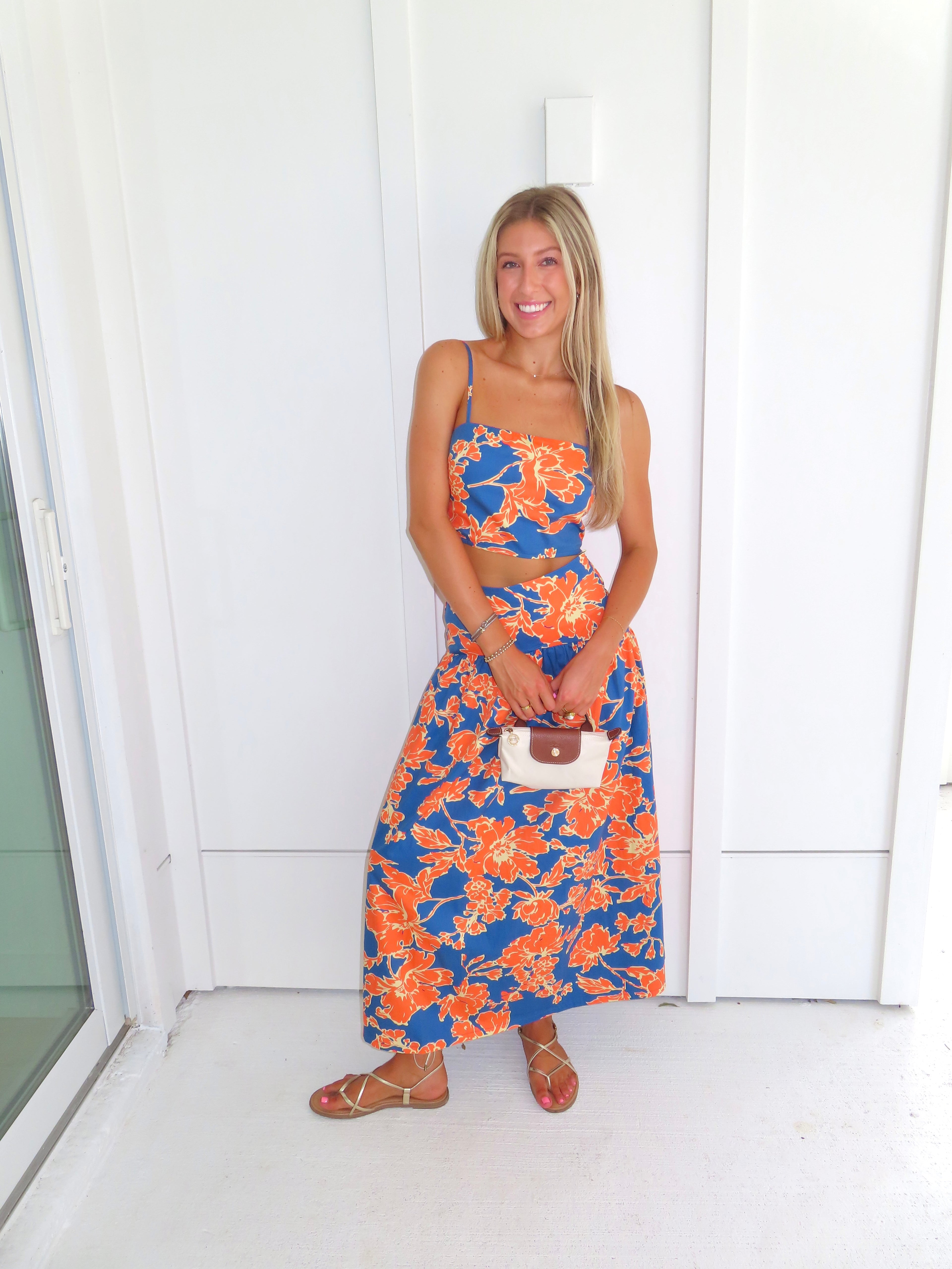 A woman posed wearing a sleeveless maxi skirt set with a bold orange and blue floral print. She is holding a small beige handbag with a brown leather detail and wearing gold strappy sandals.