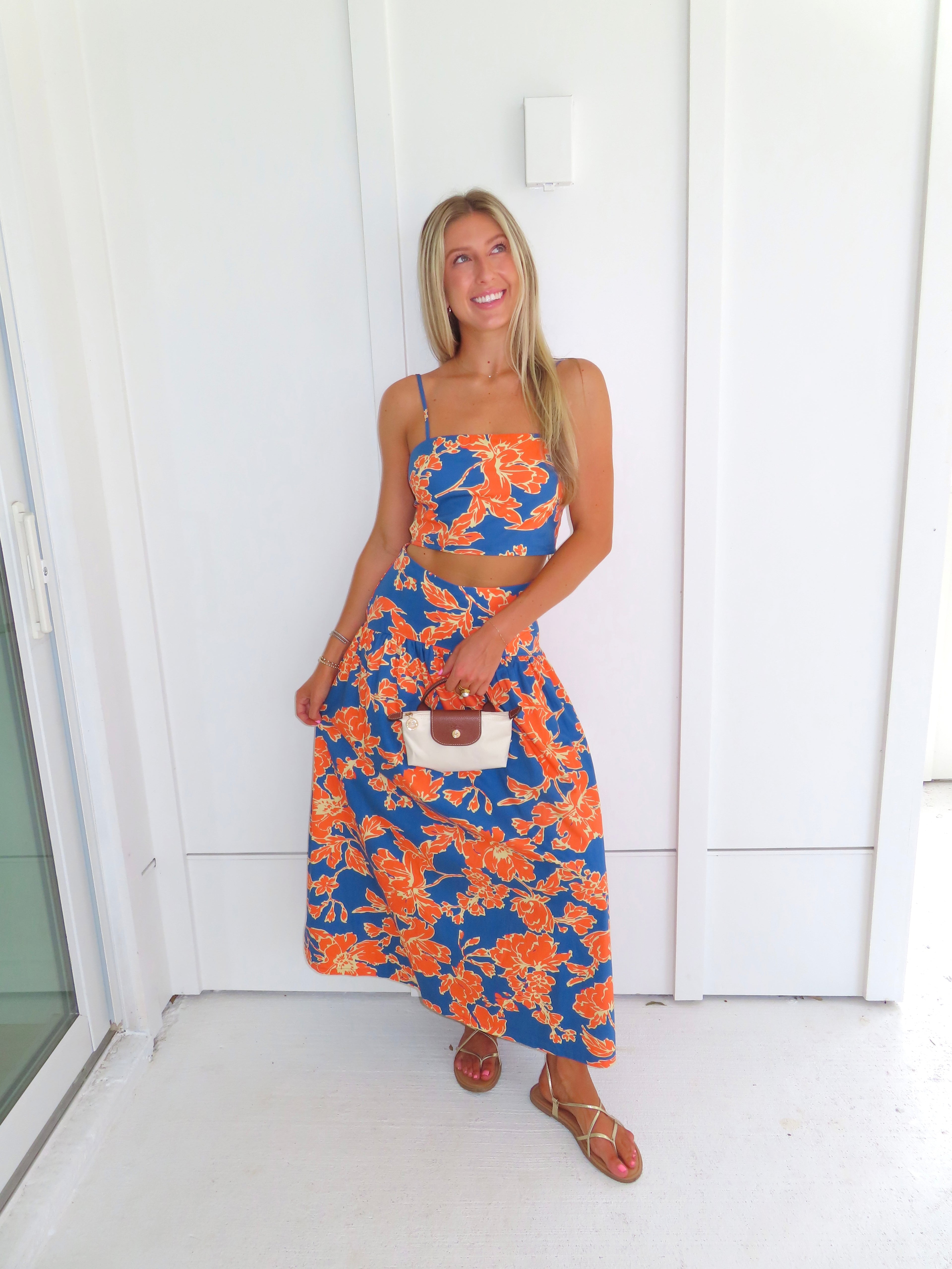 A woman posed wearing a sleeveless maxi skirt set with a bold orange and blue floral print. She is holding a small beige handbag with a brown leather detail and wearing gold strappy sandals.