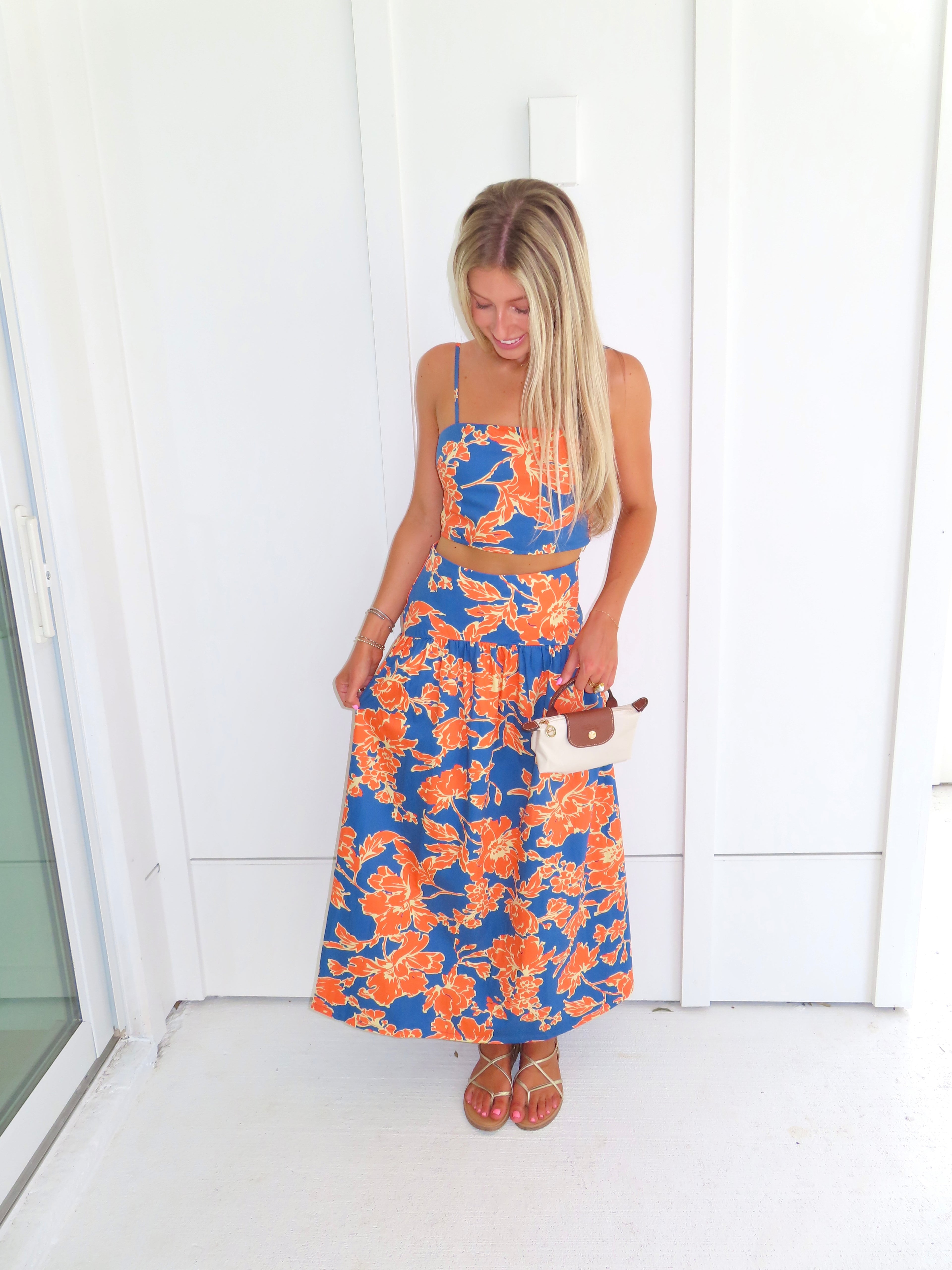 A woman posed wearing a sleeveless maxi skirt set with a bold orange and blue floral print. She is holding a small beige handbag with a brown leather detail and wearing gold strappy sandals.