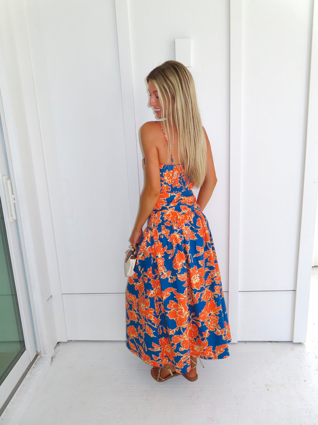 A woman posed wearing a sleeveless maxi skirt set with a bold orange and blue floral print. She is holding a small beige handbag with a brown leather detail and wearing gold strappy sandals.