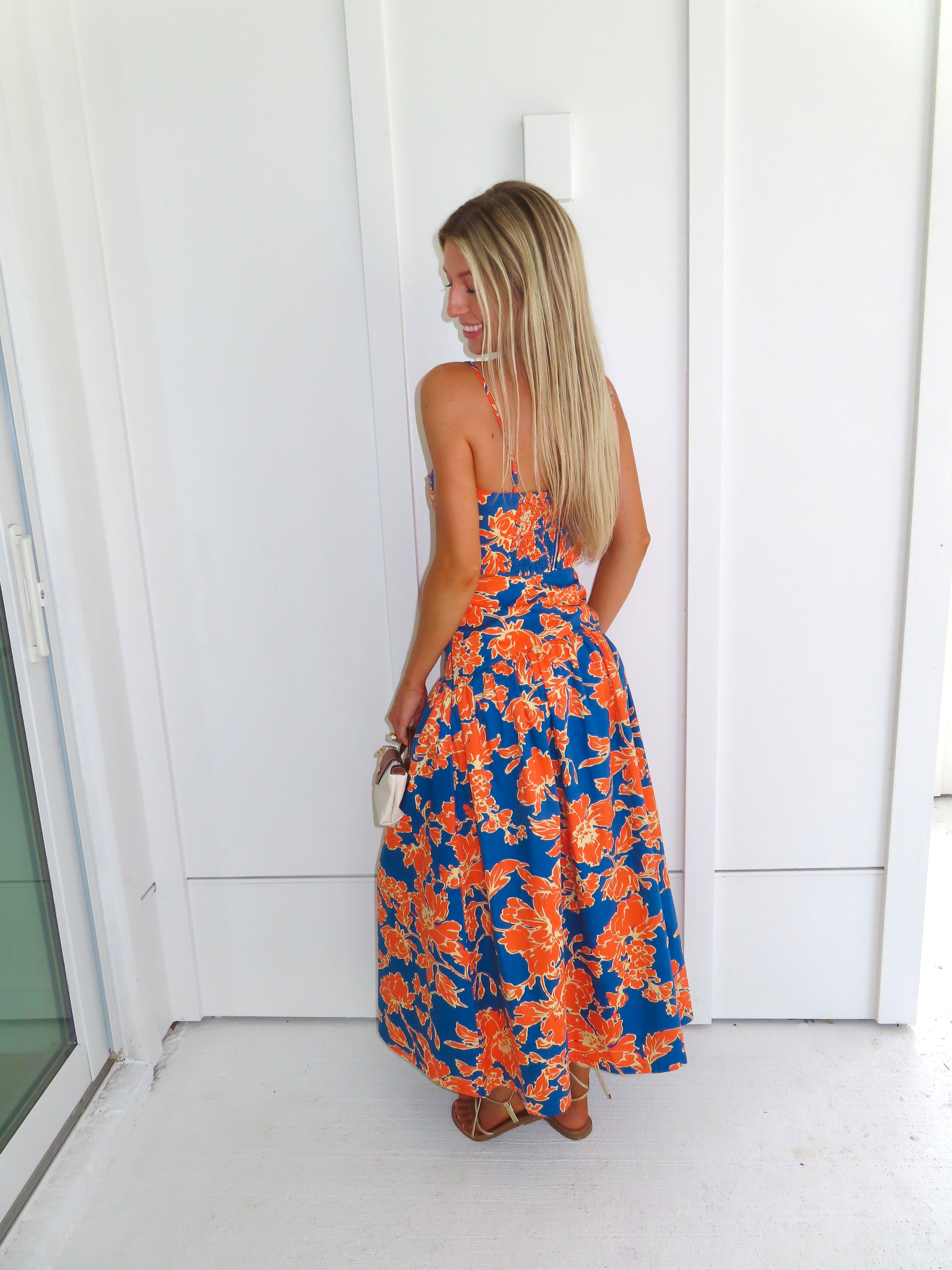 A woman posed wearing a sleeveless maxi skirt set with a bold orange and blue floral print. She is holding a small beige handbag with a brown leather detail and wearing gold strappy sandals.