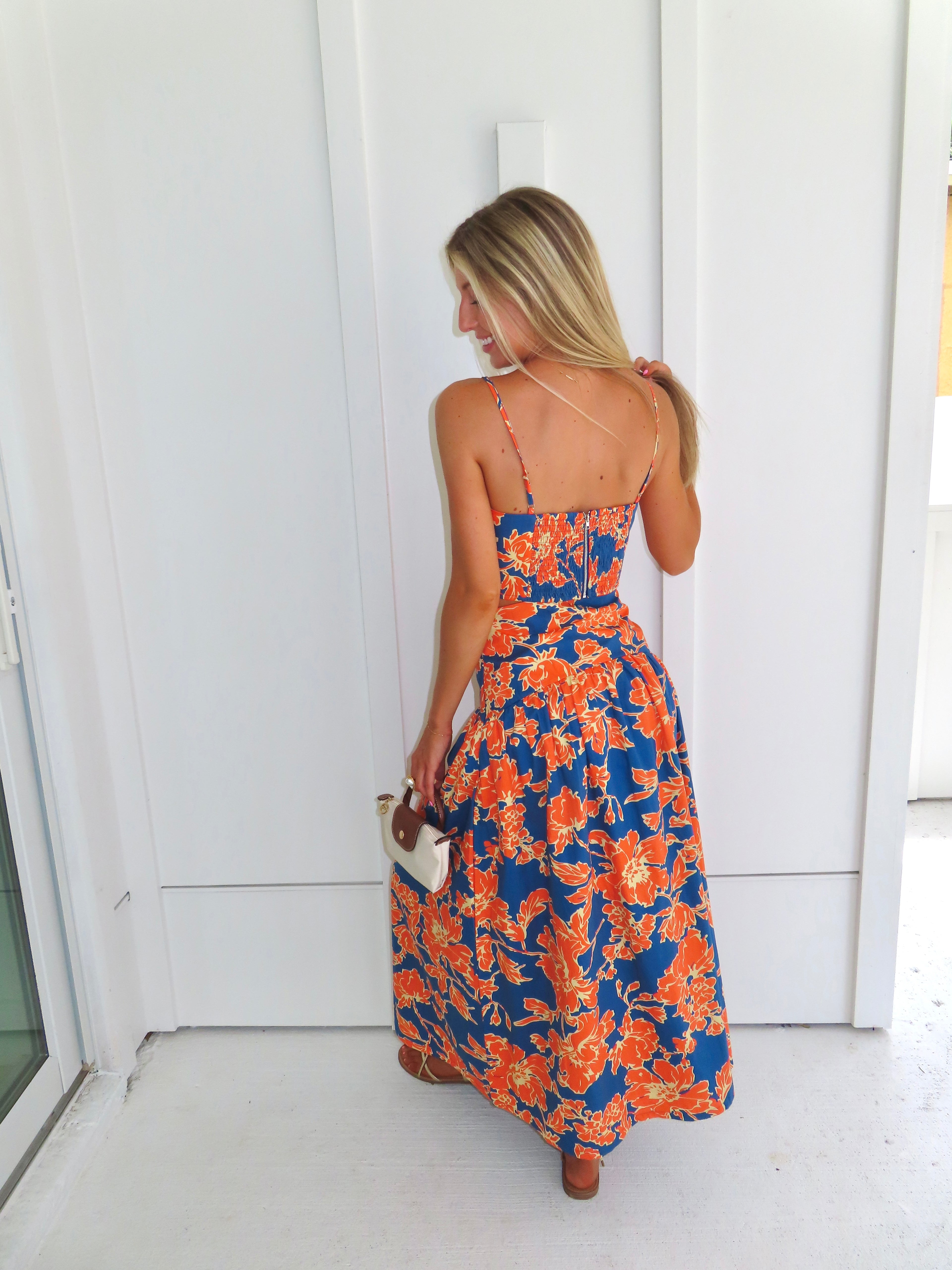 A woman posed wearing a sleeveless maxi skirt set with a bold orange and blue floral print. She is holding a small beige handbag with a brown leather detail and wearing gold strappy sandals.