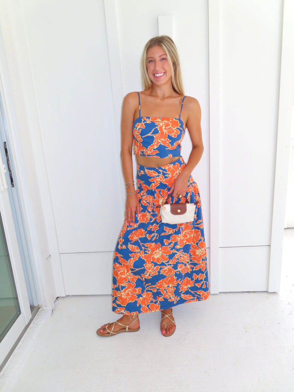 A woman posed wearing a sleeveless maxi skirt set with a bold orange and blue floral print. She is holding a small beige handbag with a brown leather detail and wearing gold strappy sandals.