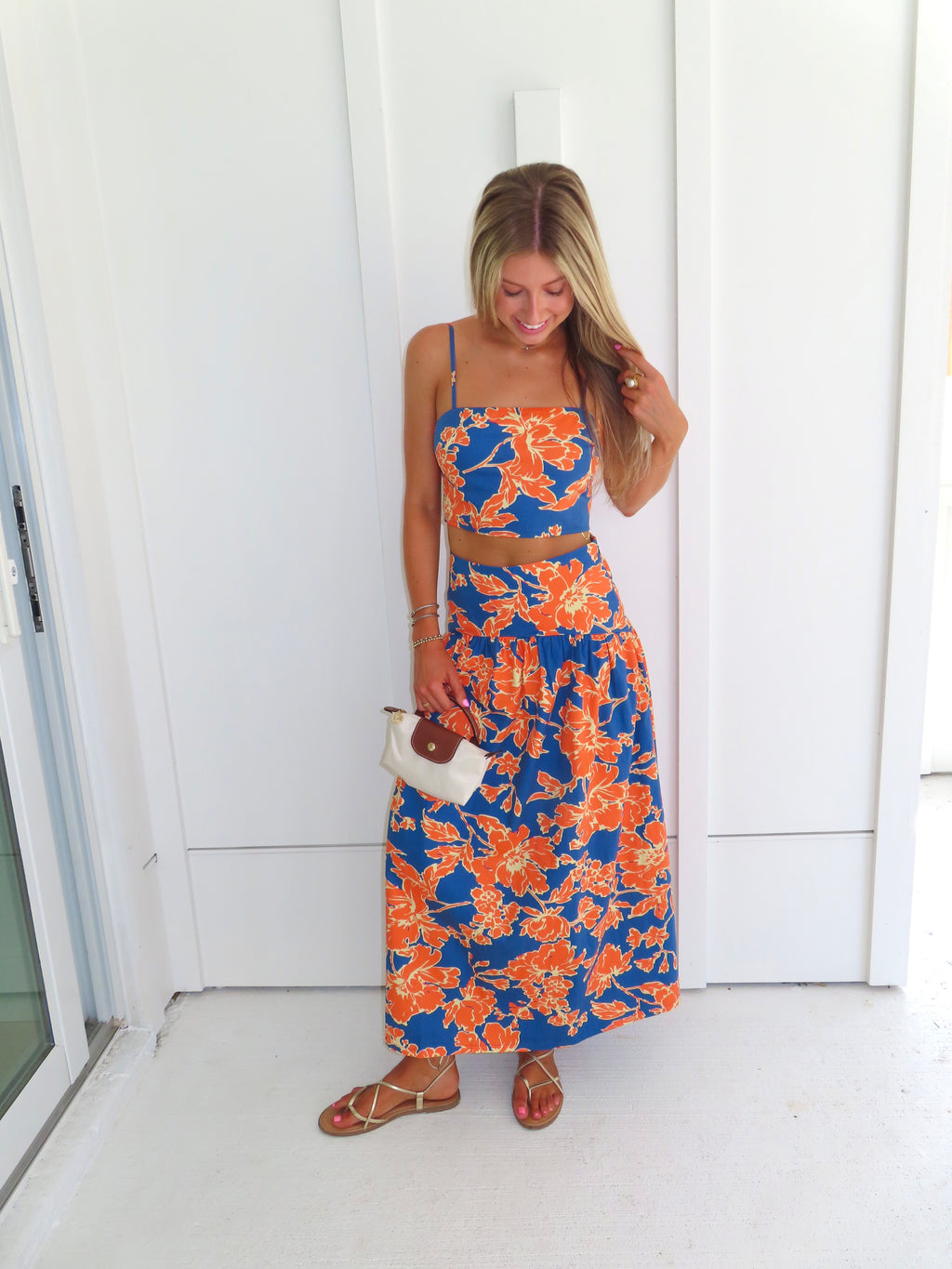 A woman posed wearing a sleeveless maxi skirt set with a bold orange and blue floral print. She is holding a small beige handbag with a brown leather detail and wearing gold strappy sandals.