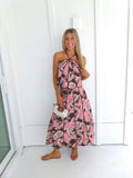 Woman standing in front of a white paneled wall wearing a floral pink and brown tank top and maxi skirt set, holding a beige purse and wearing gold sandals.