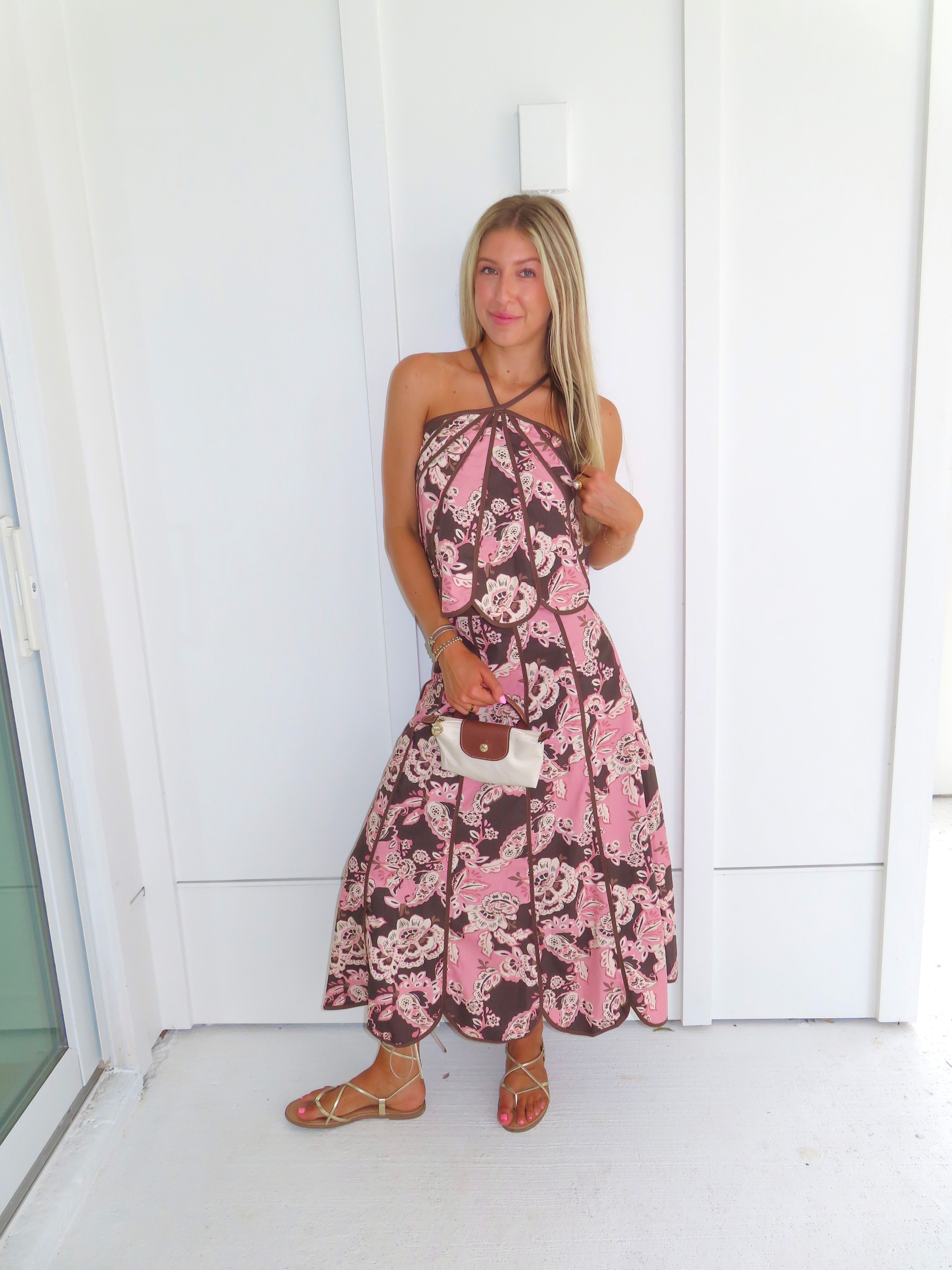 Woman standing in front of a white paneled wall wearing a floral pink and brown tank top and maxi skirt set, holding a beige purse and wearing gold sandals.