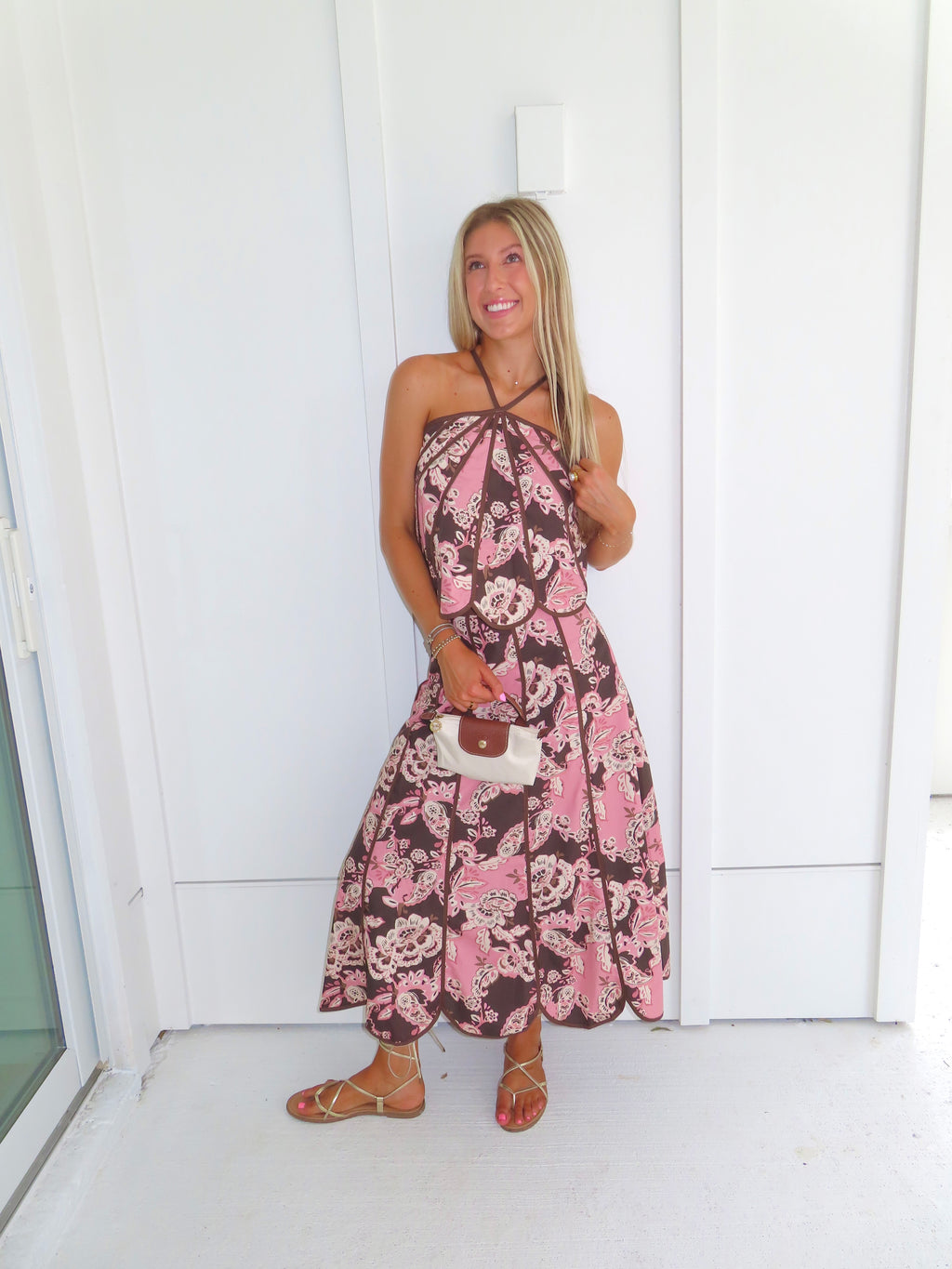 Woman standing in front of a white paneled wall wearing a floral pink and brown tank top and maxi skirt set, holding a beige purse and wearing gold sandals.