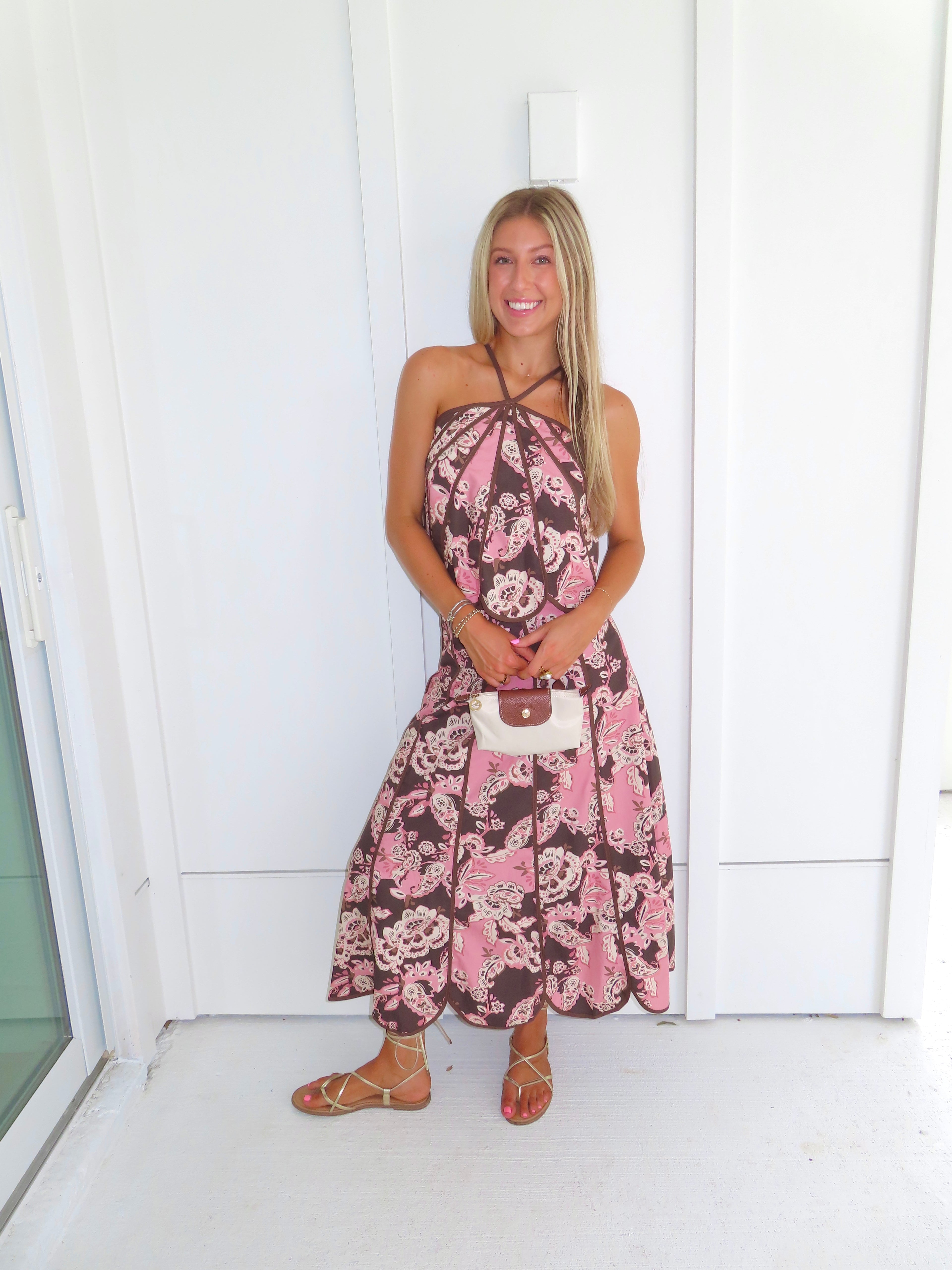 Woman standing in front of a white paneled wall wearing a floral pink and brown tank top and maxi skirt set, holding a beige purse and wearing gold sandals.