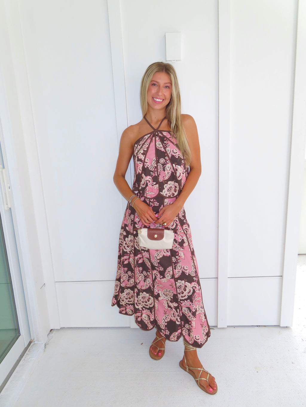 Woman standing in front of a white paneled wall wearing a floral pink and brown tank top and maxi skirt set, holding a beige purse and wearing gold sandals.