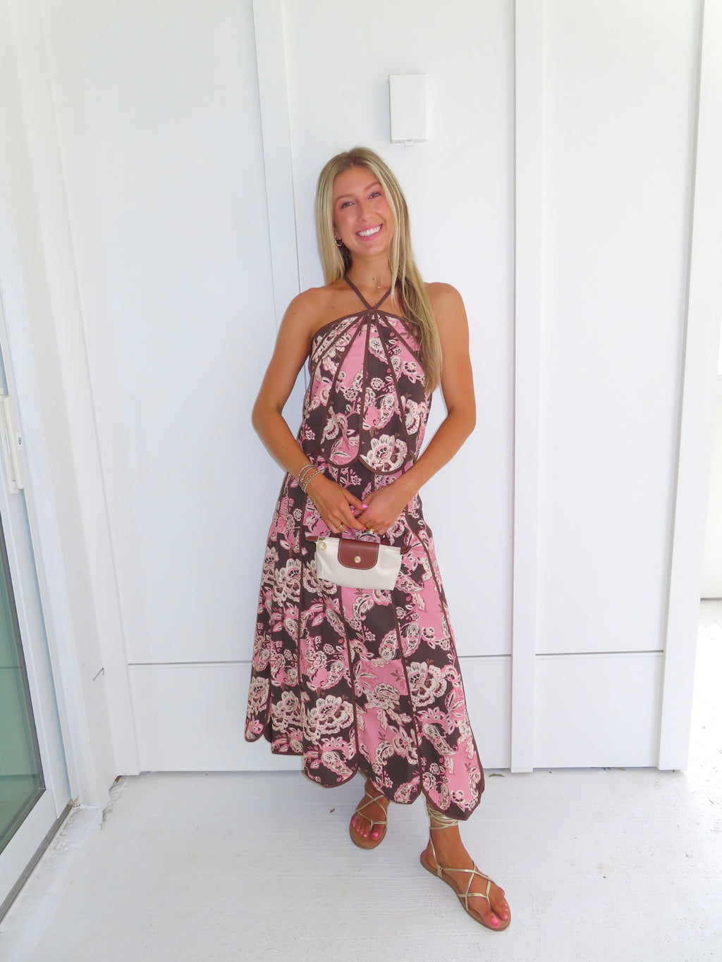 Woman standing in front of a white paneled wall wearing a floral pink and brown tank top and maxi skirt set, holding a beige purse and wearing gold sandals.