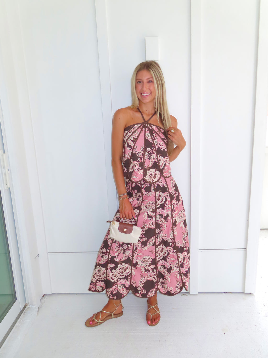 Woman standing in front of a white paneled wall wearing a floral pink and brown tank top and maxi skirt set, holding a beige purse and wearing gold sandals.