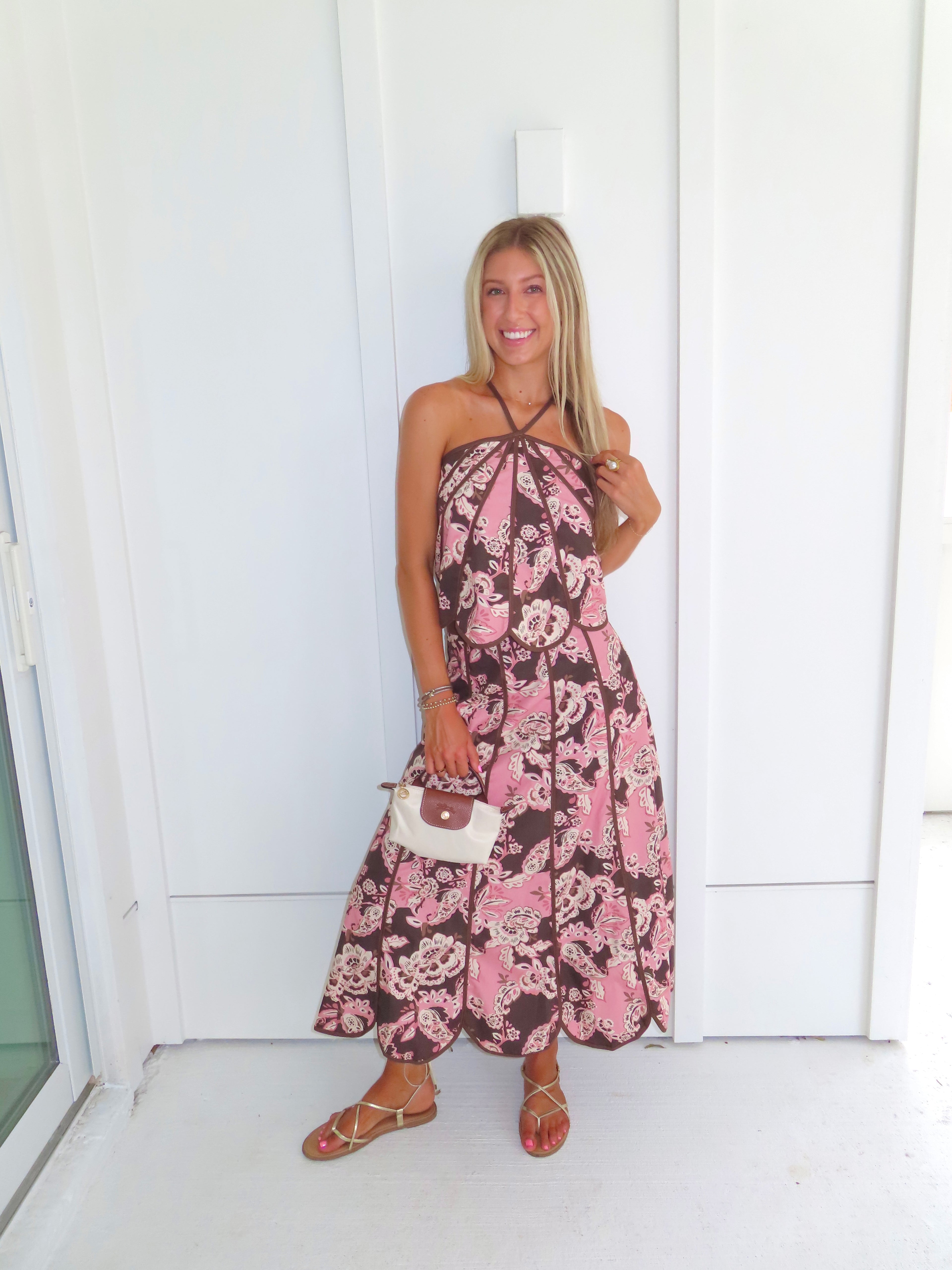 Woman standing in front of a white paneled wall wearing a floral pink and brown tank top and maxi skirt set, holding a beige purse and wearing gold sandals.