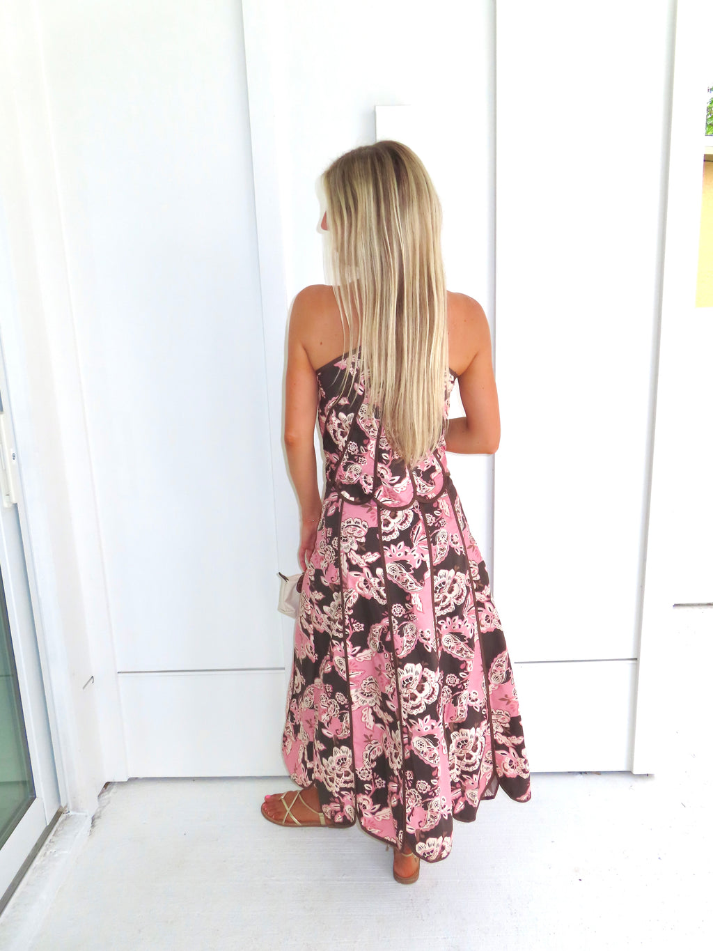 Woman standing in front of a white paneled wall wearing a floral pink and brown tank top and maxi skirt set, holding a beige purse and wearing gold sandals.