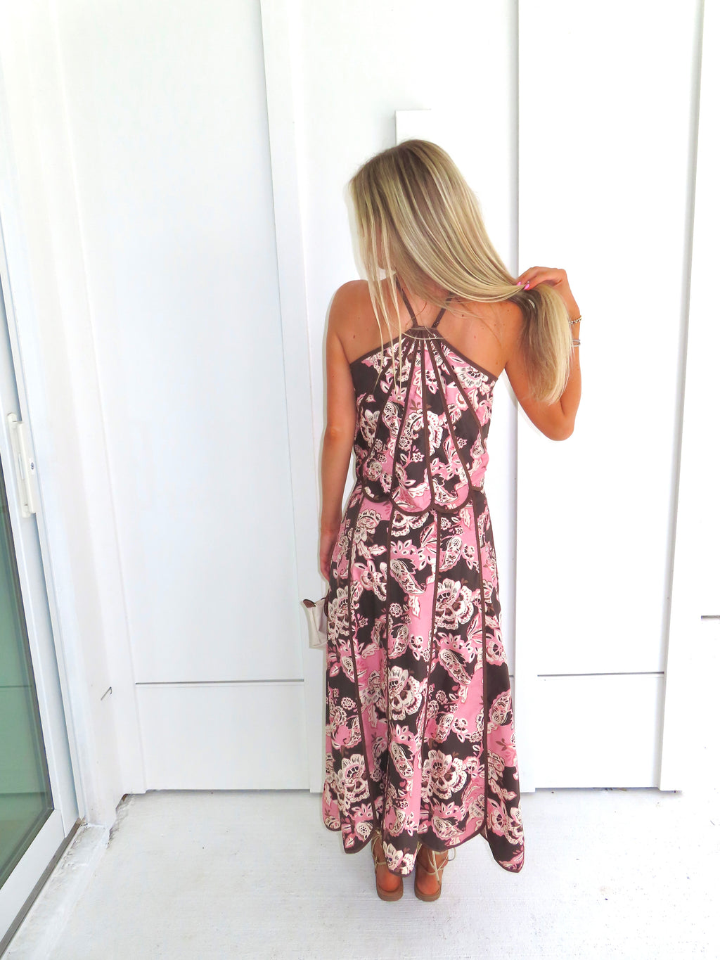 Woman standing in front of a white paneled wall wearing a floral pink and brown tank top and maxi skirt set, holding a beige purse and wearing gold sandals.
