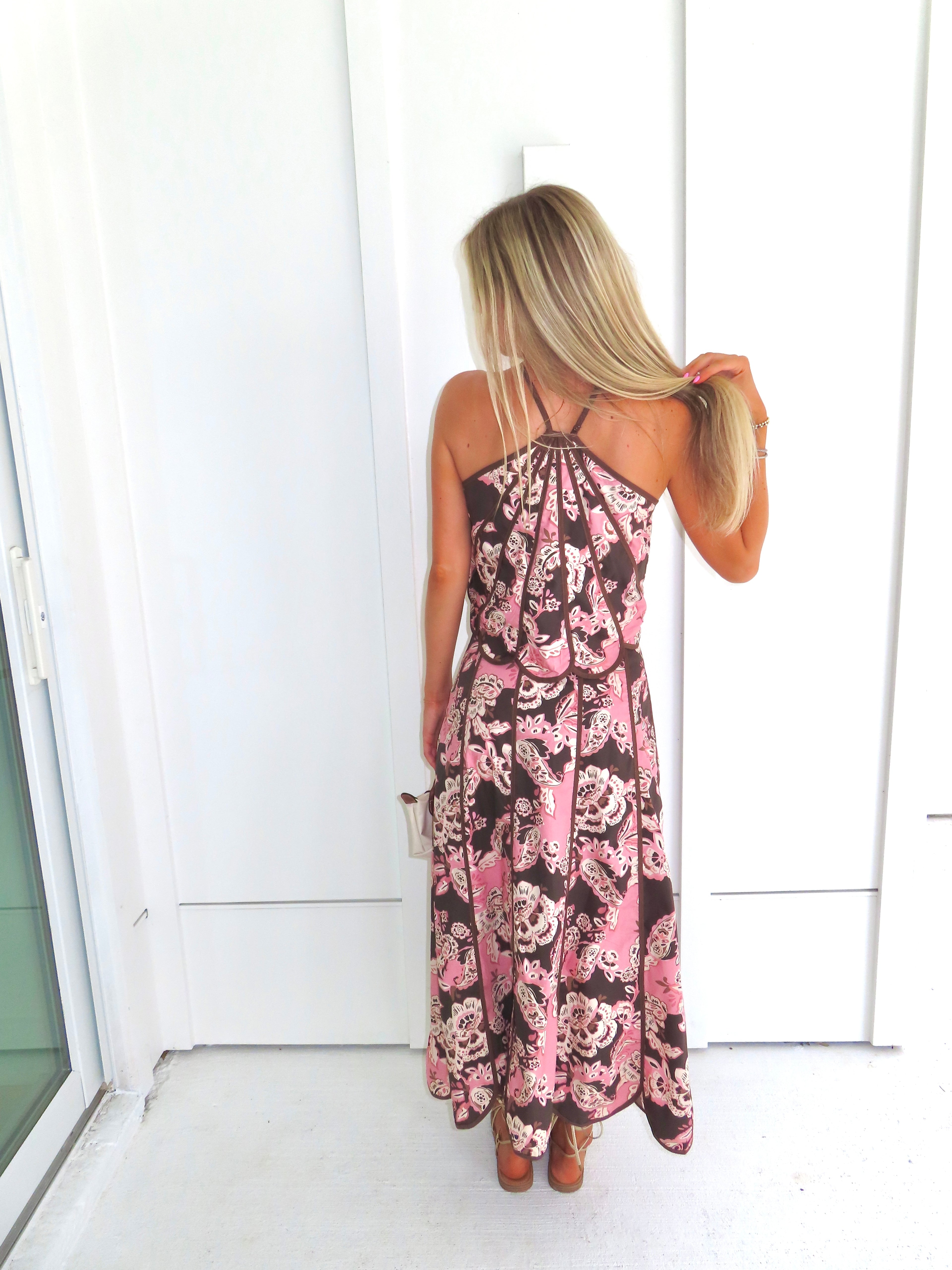 Woman standing in front of a white paneled wall wearing a floral pink and brown tank top and maxi skirt set, holding a beige purse and wearing gold sandals.