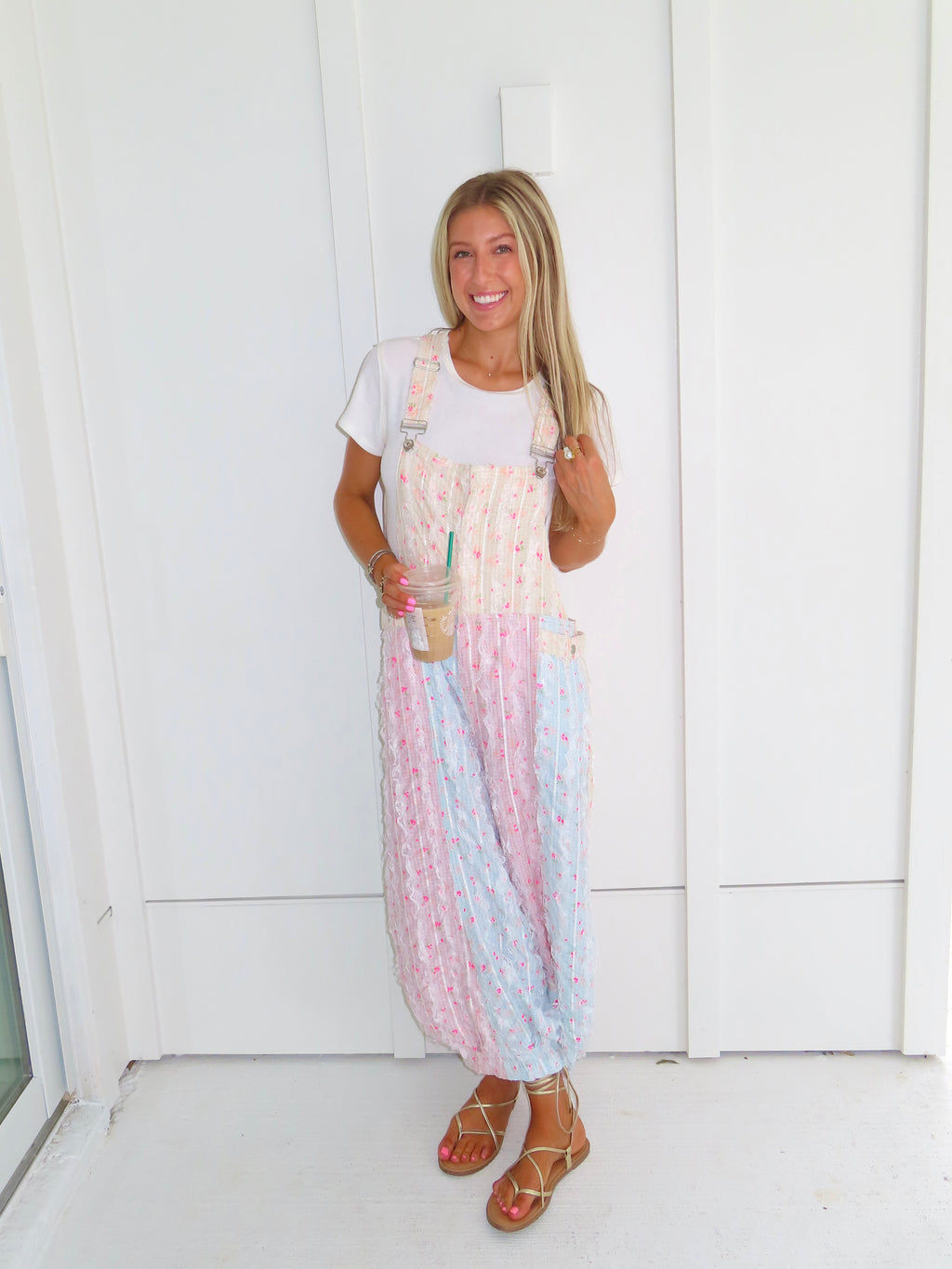 A woman wearing a pastel patchwork overall jumpsuit with a white t-shirt underneath and gold sandals, holding a coffee and standing against a white paneled wall.