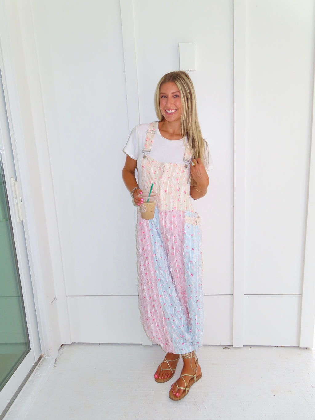 A woman wearing a pastel patchwork overall jumpsuit with a white t-shirt underneath and gold sandals, holding a coffee and standing against a white paneled wall.