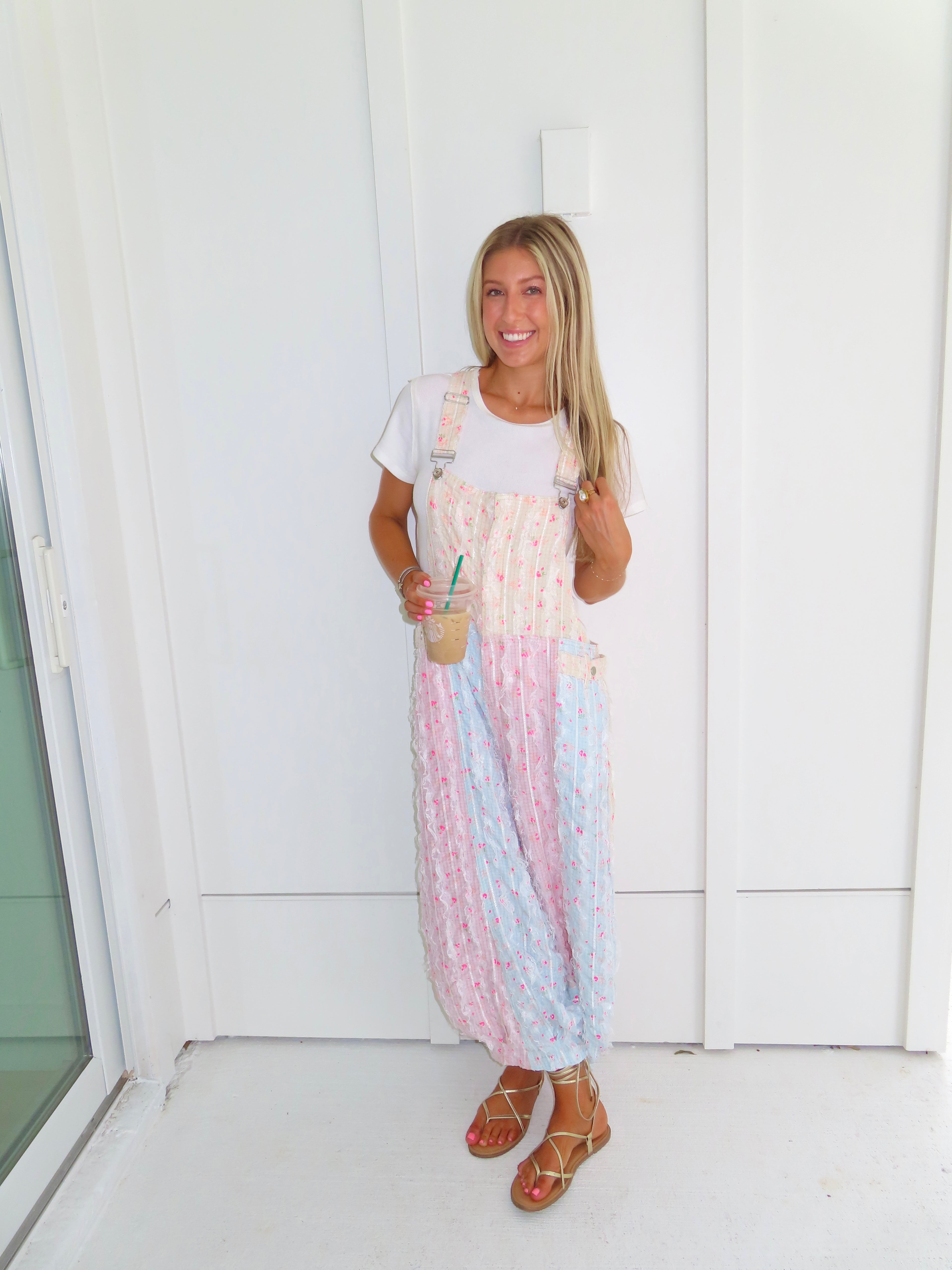 A woman wearing a pastel patchwork overall jumpsuit with a white t-shirt underneath and gold sandals, holding a coffee and standing against a white paneled wall.