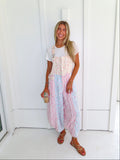 A woman wearing a pastel patchwork overall jumpsuit with a white t-shirt underneath and gold sandals, holding a coffee and standing against a white paneled wall.