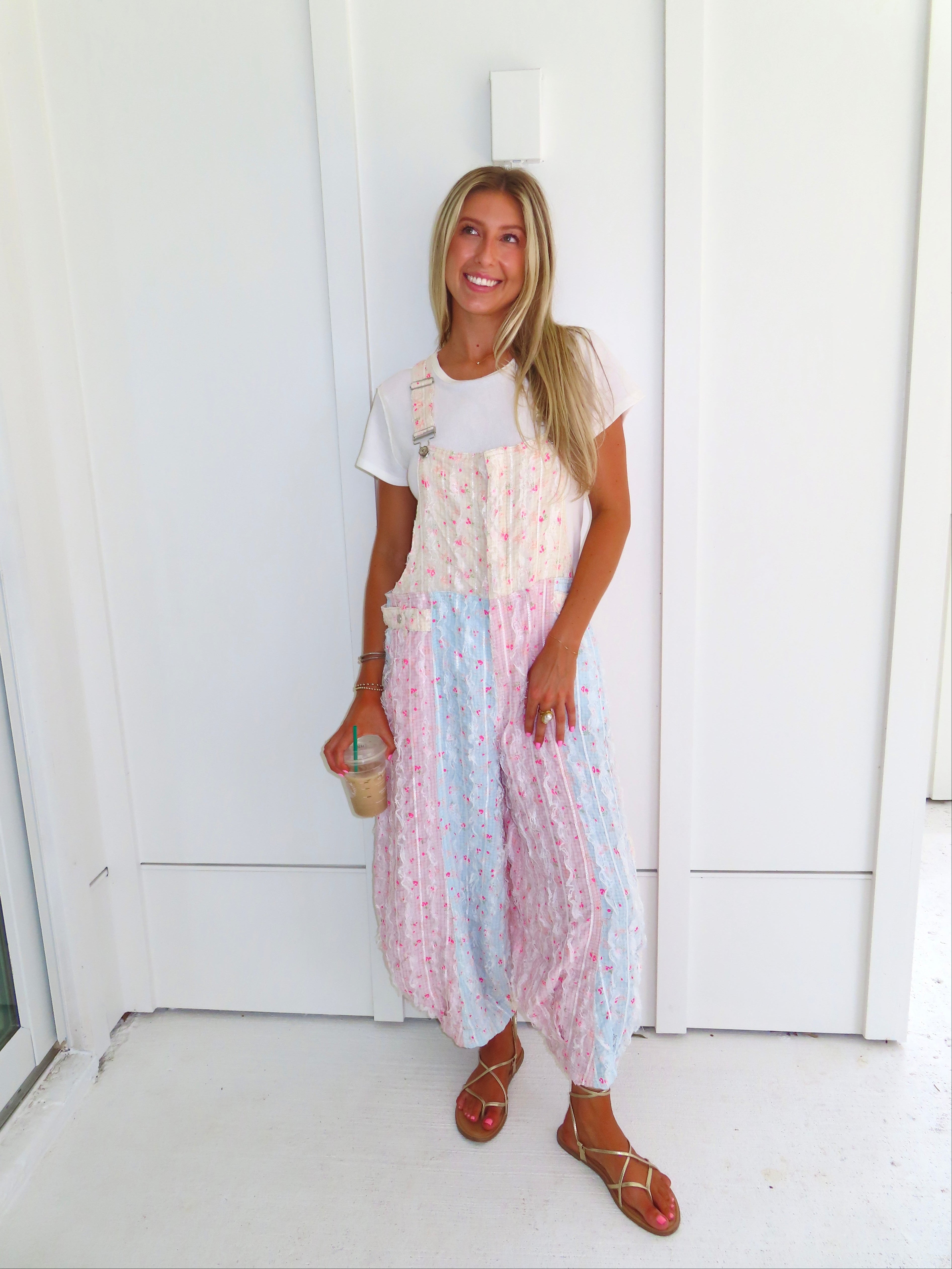 A woman wearing a pastel patchwork overall jumpsuit with a white t-shirt underneath and gold sandals, holding a coffee and standing against a white paneled wall.