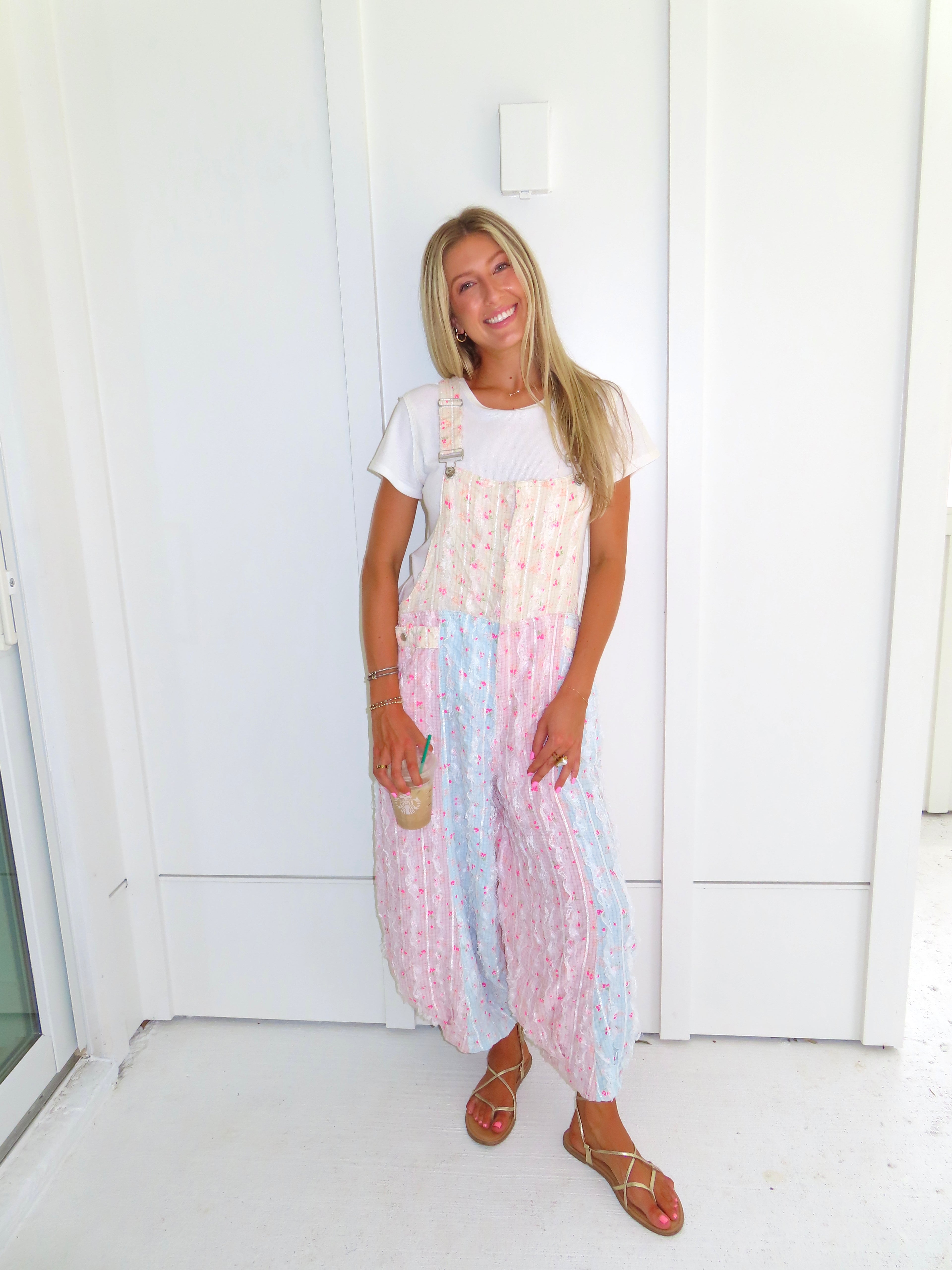 A woman wearing a pastel patchwork overall jumpsuit with a white t-shirt underneath and gold sandals, holding a coffee and standing against a white paneled wall.