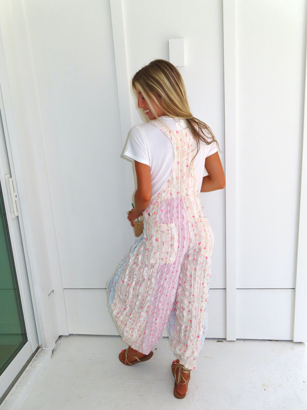 A woman wearing a pastel patchwork overall jumpsuit with a white t-shirt underneath and gold sandals, holding a coffee and standing against a white paneled wall.