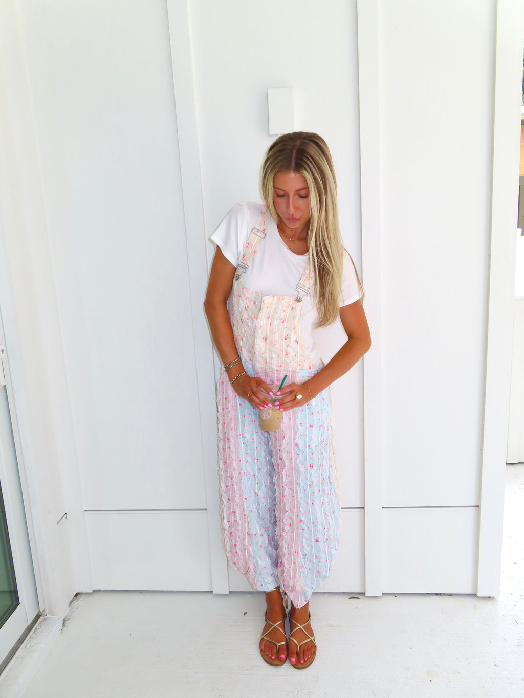 A woman wearing a pastel patchwork overall jumpsuit with a white t-shirt underneath and gold sandals, holding a coffee and standing against a white paneled wall.