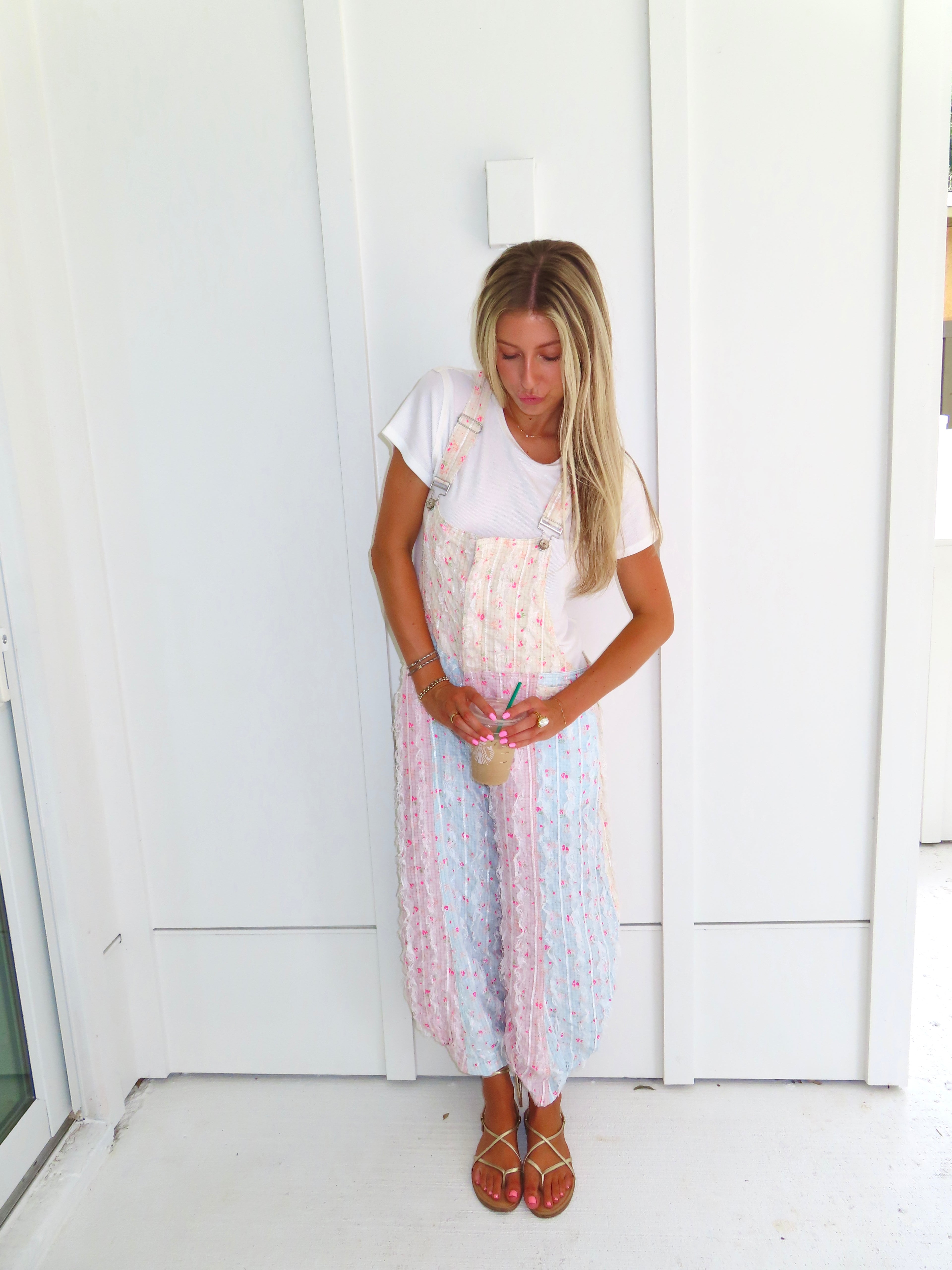 A woman wearing a pastel patchwork overall jumpsuit with a white t-shirt underneath and gold sandals, holding a coffee and standing against a white paneled wall.