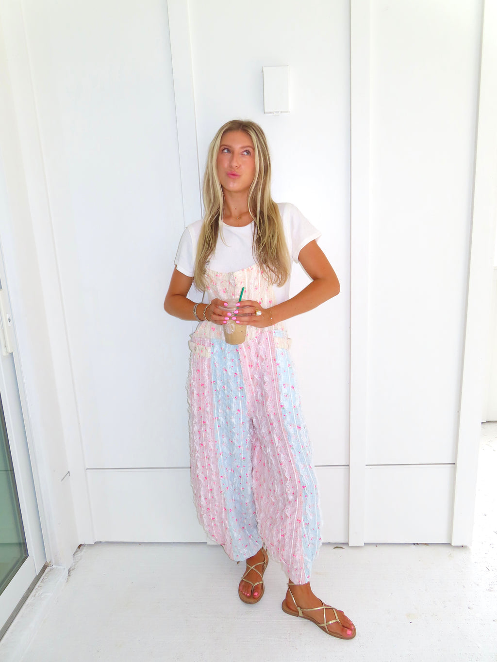 A woman wearing a pastel patchwork overall jumpsuit with a white t-shirt underneath and gold sandals, holding a coffee and standing against a white paneled wall.