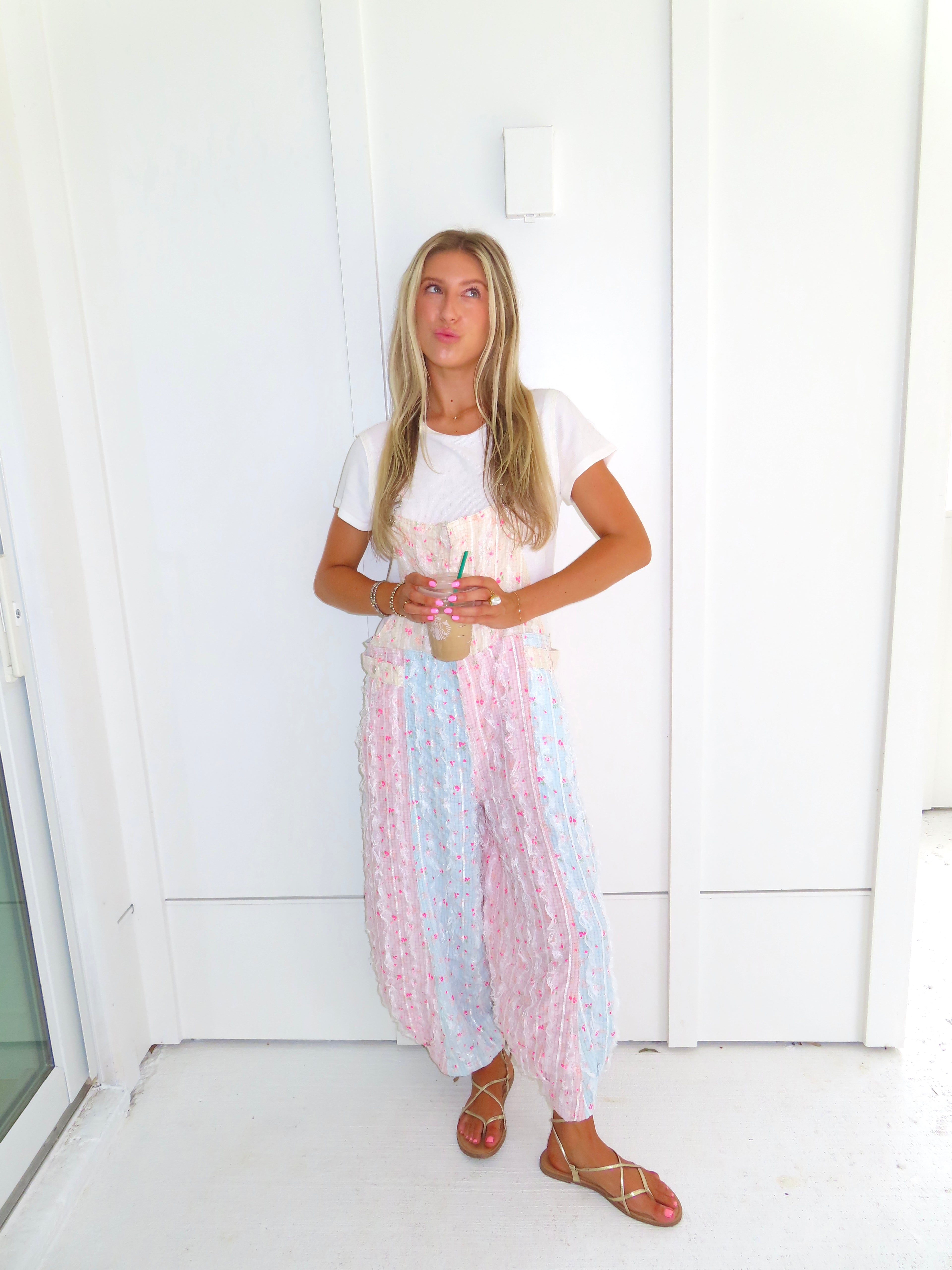 A woman wearing a pastel patchwork overall jumpsuit with a white t-shirt underneath and gold sandals, holding a coffee and standing against a white paneled wall.