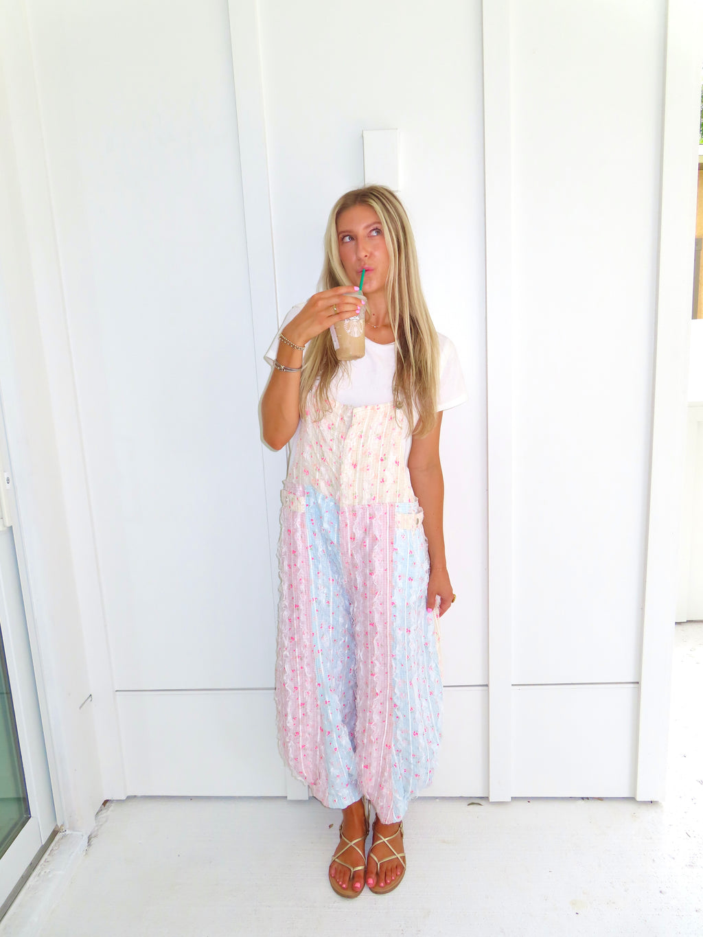A woman wearing a pastel patchwork overall jumpsuit with a white t-shirt underneath and gold sandals, holding a coffee and standing against a white paneled wall.