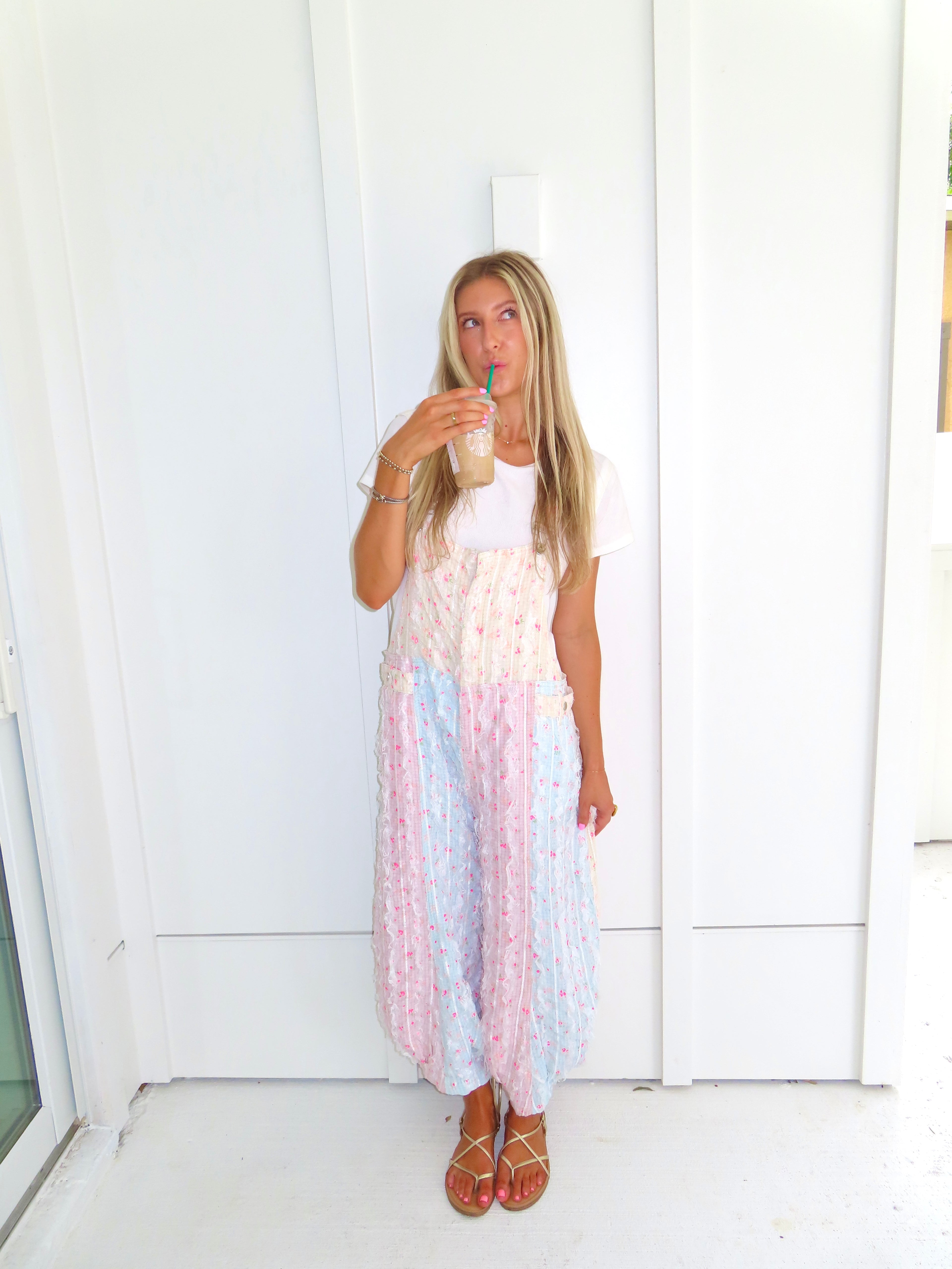 A woman wearing a pastel patchwork overall jumpsuit with a white t-shirt underneath and gold sandals, holding a coffee and standing against a white paneled wall.