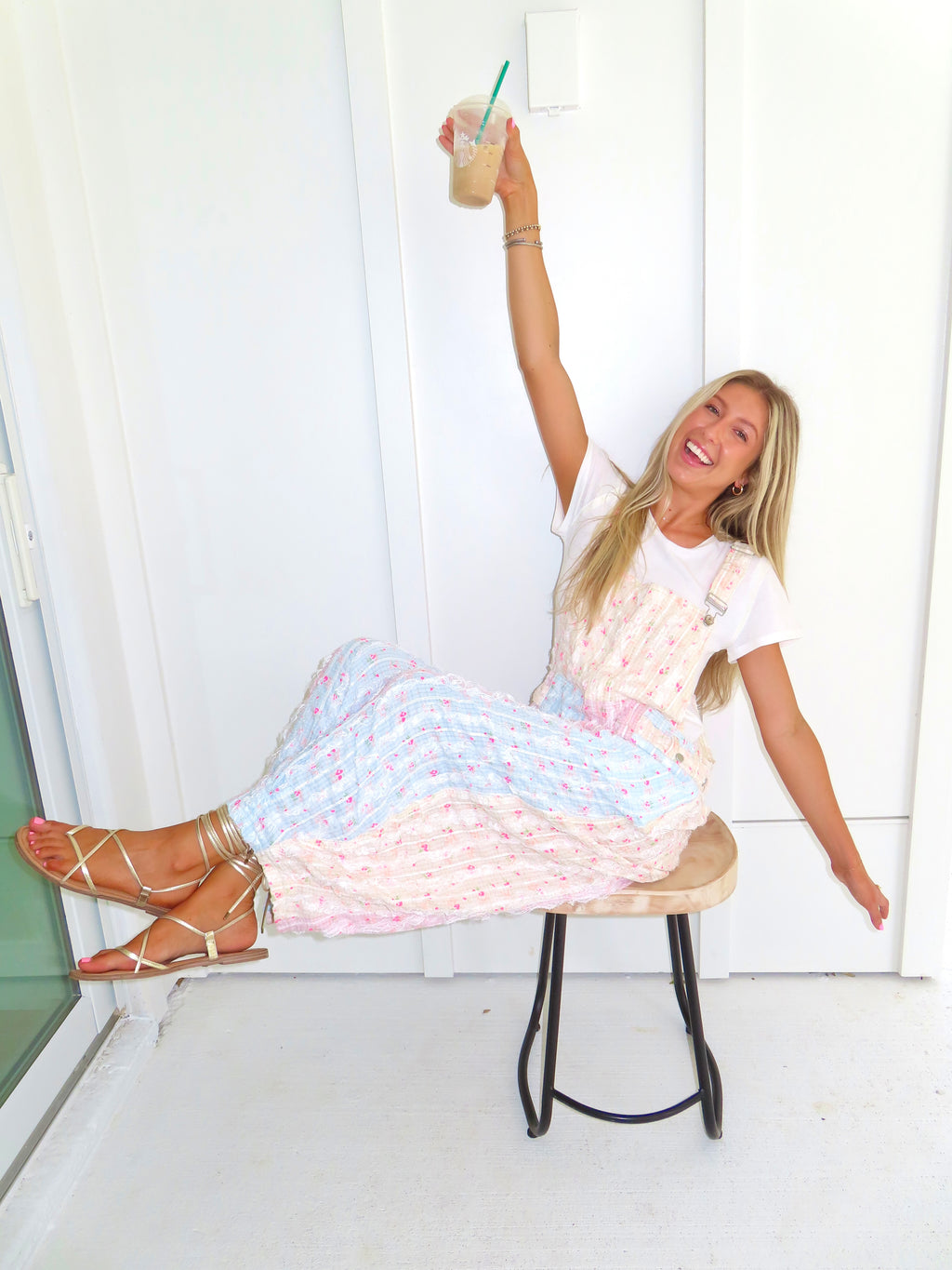 A woman sitting on a stool wearing a white t-shirt and pastel pink, blue, and yellow patterned overalls and gold sandals. She is holding up a iced coffee with a green straw.