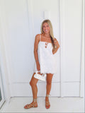 A woman stands against a white paneled wall wearing a white spaghetti-strap mini dress with a lace-up cutout at the bust, holding a small white handbag and wearing tan lace-up sandals.