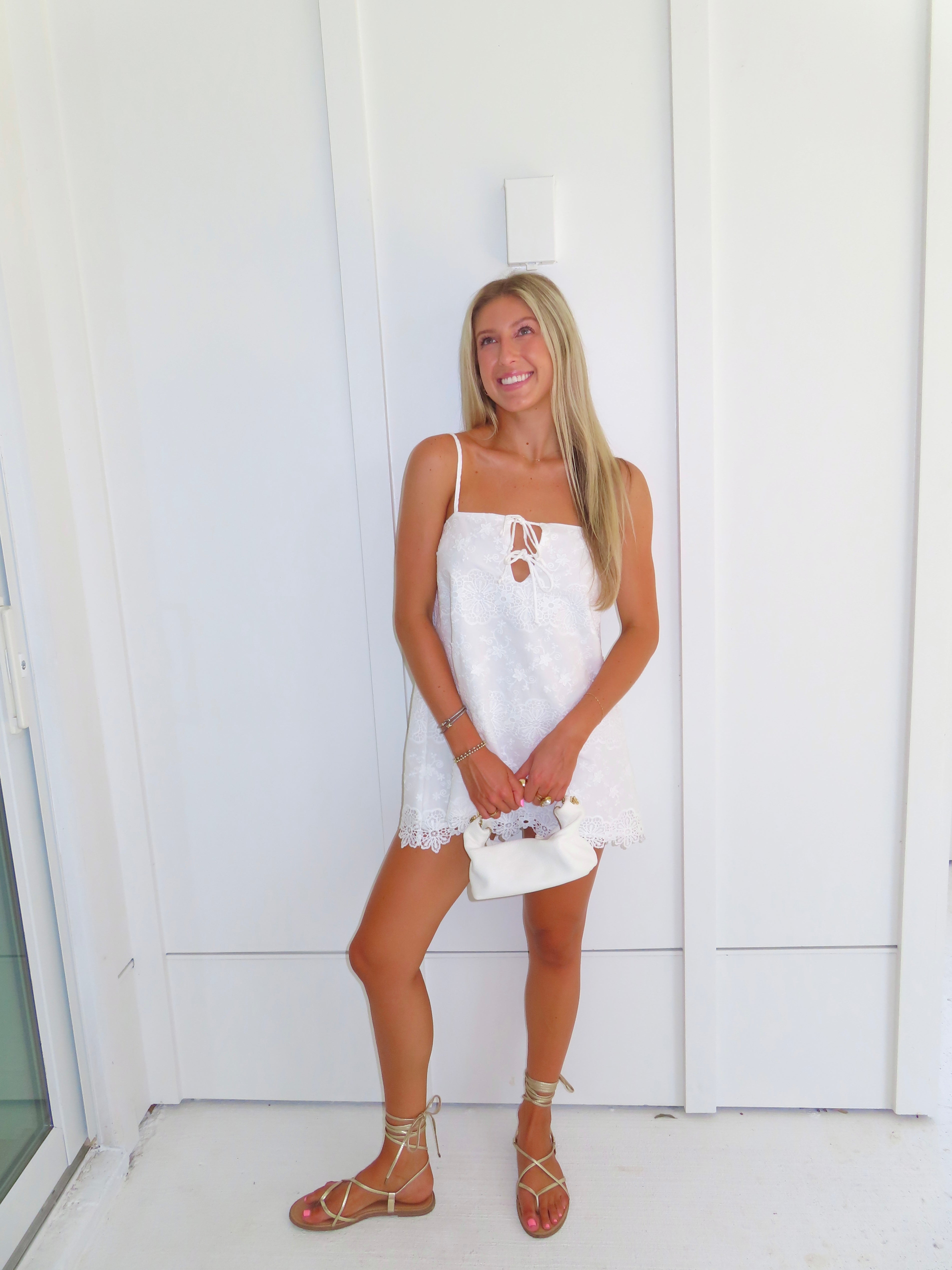 A woman stands against a white paneled wall wearing a white spaghetti-strap mini dress with a lace-up cutout at the bust, holding a small white handbag and wearing tan lace-up sandals.