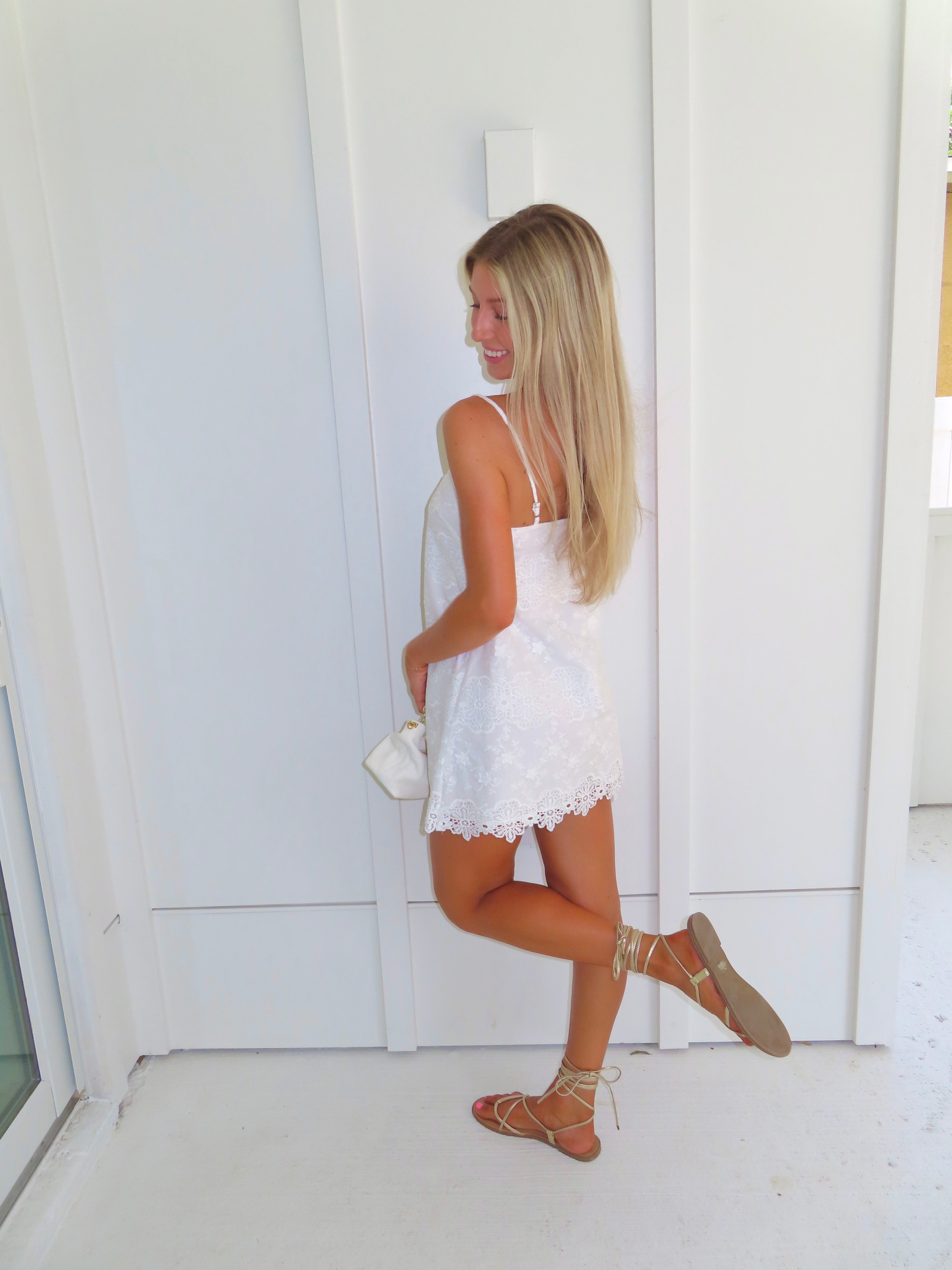 A woman stands against a white paneled wall wearing a white spaghetti-strap mini dress with a lace-up cutout at the bust, holding a small white handbag and wearing tan lace-up sandals.