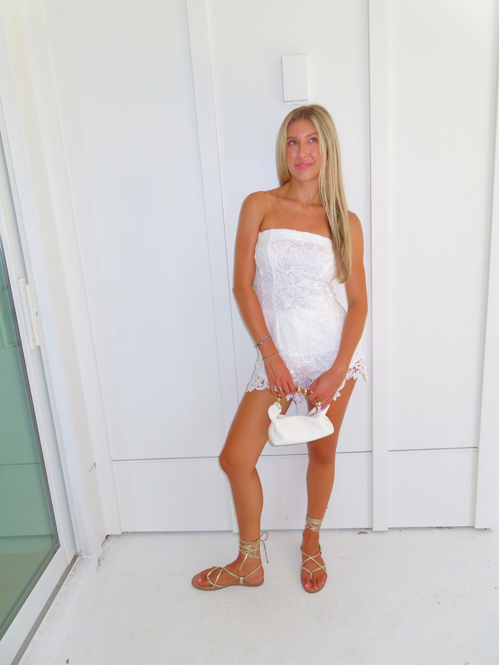 A woman posing in a white strapless lace strapless top and shorts set, holding a small white handbag and wearing gold strappy flat sandals, standing against a white paneled wall.