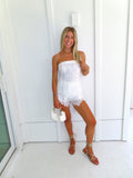 A woman posing in a white strapless lace strapless top and shorts set, holding a small white handbag and wearing gold strappy flat sandals, standing against a white paneled wall.