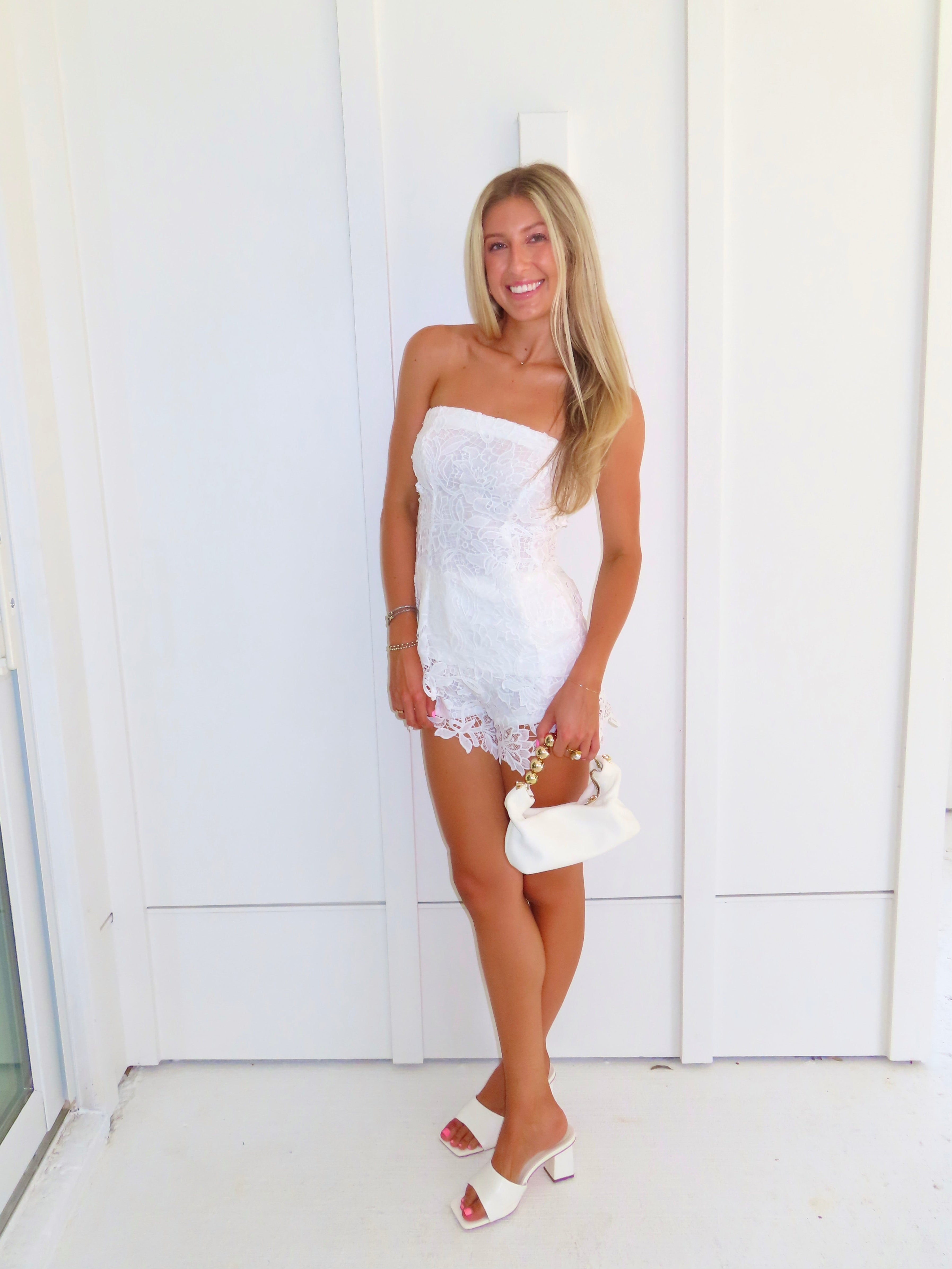 A woman posing in a white strapless lace strapless top and shorts set, holding a small white handbag and wearing gold strappy flat sandals, standing against a white paneled wall.